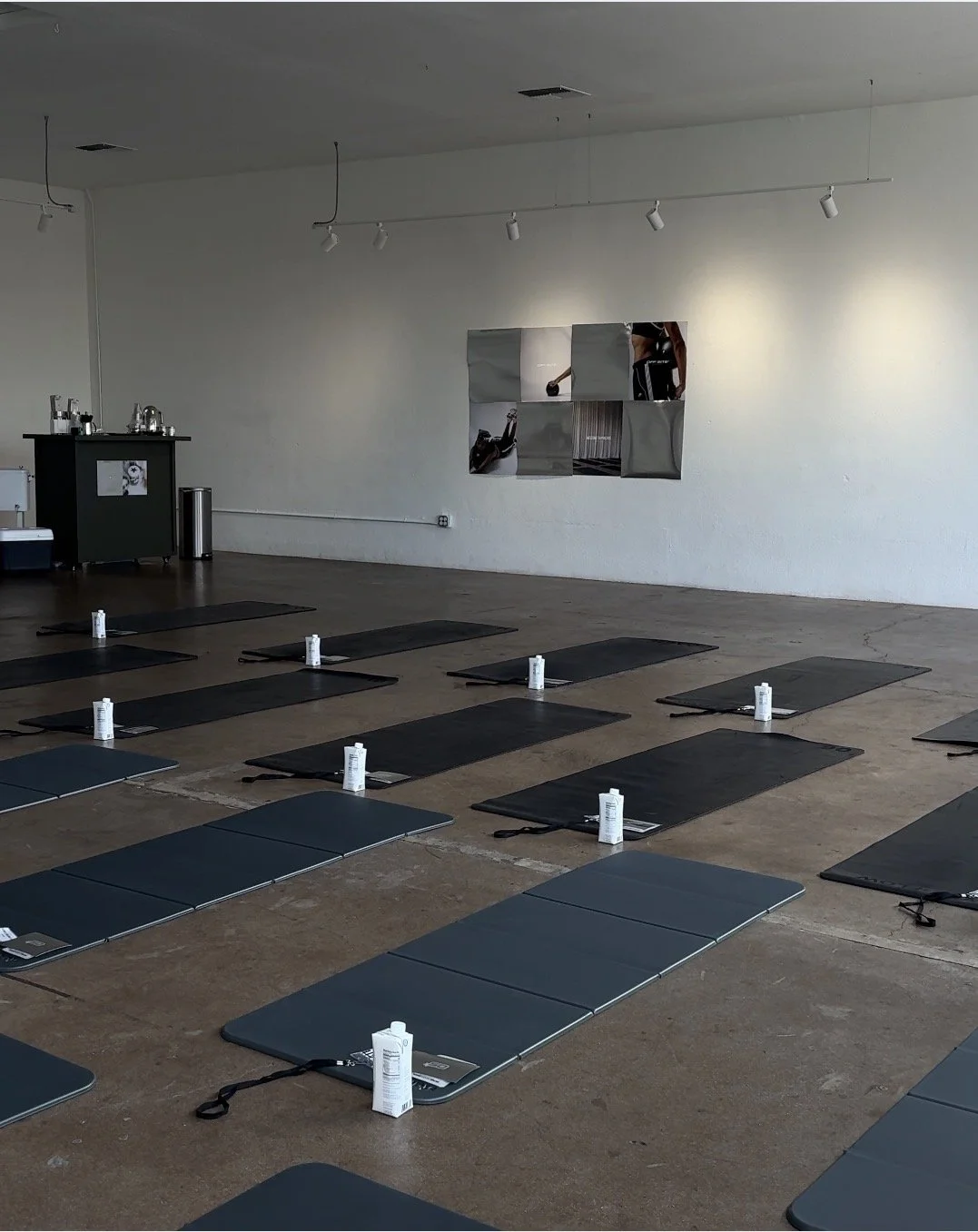 Indoor yoga or fitness class setup with black mats, bottled water, and white towels on a wooden floor in a room with white walls and track lighting.