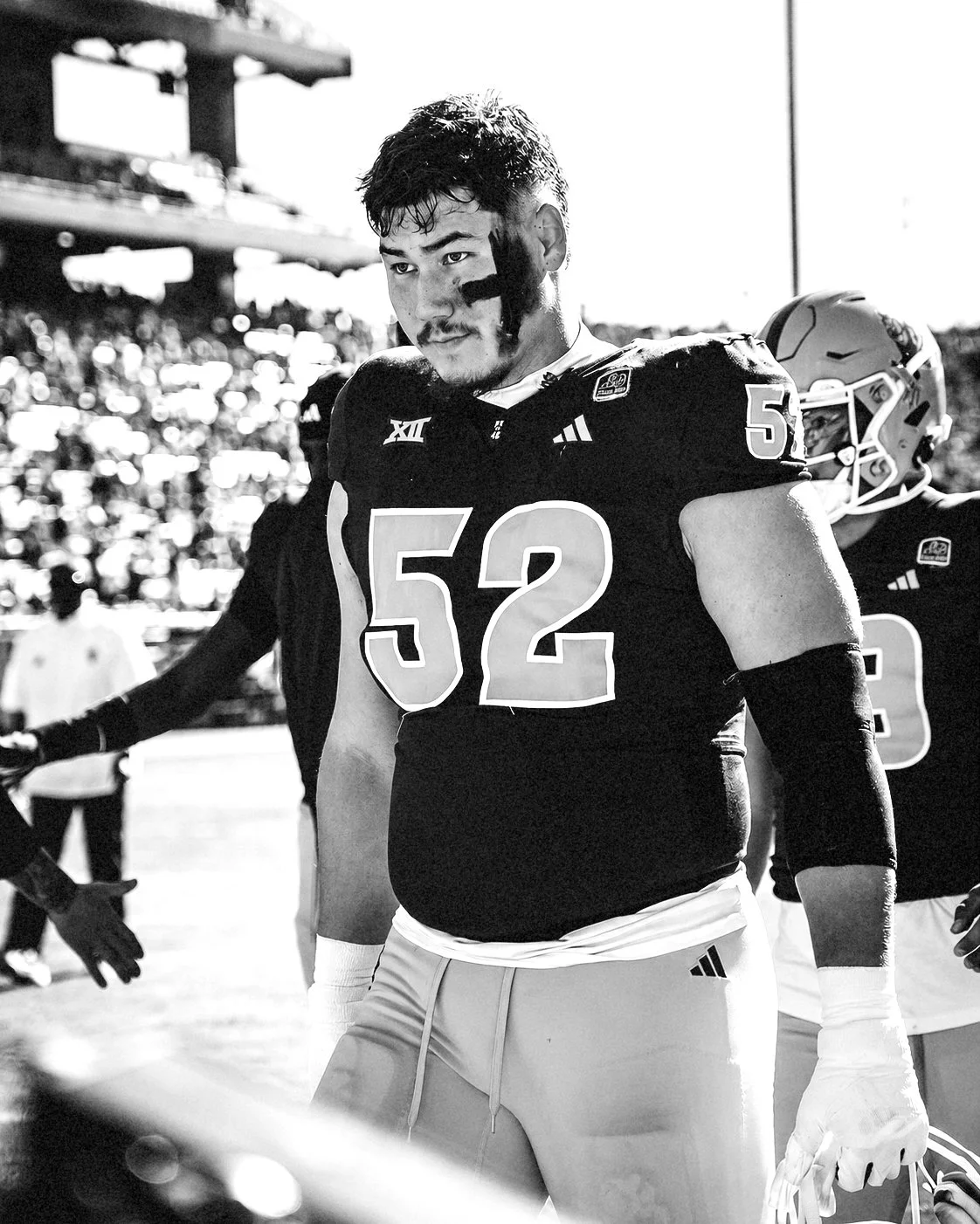 A football player wearing a jersey with the number 52, standing on the sideline during a game, with a face paint stripe across his face and a serious expression.