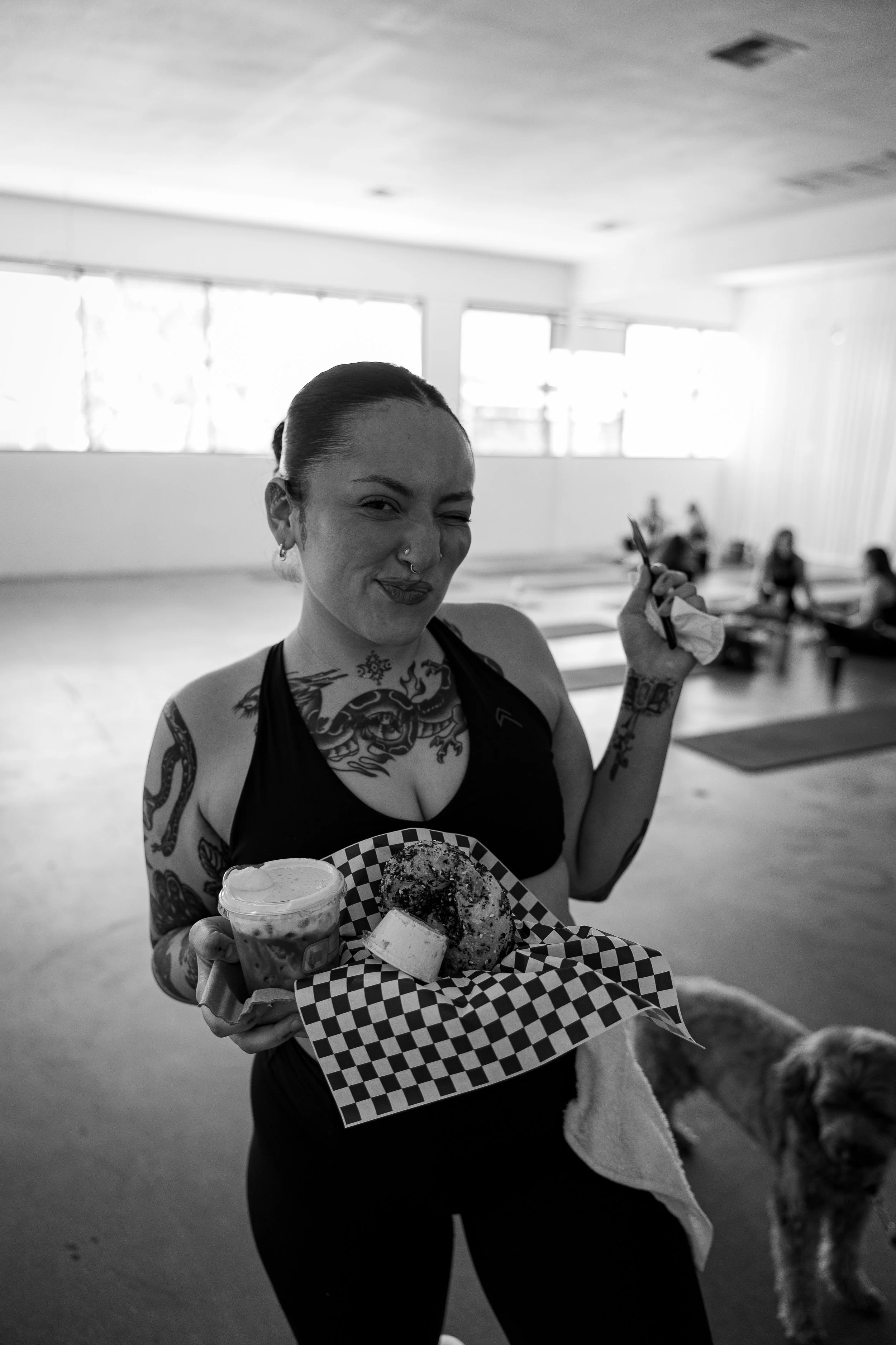 A woman with tattoos, piercings, and a nose ring, smiling and winking while holding a tray with a donut and a drink, and a white towel in her other hand, in a bright indoor space.