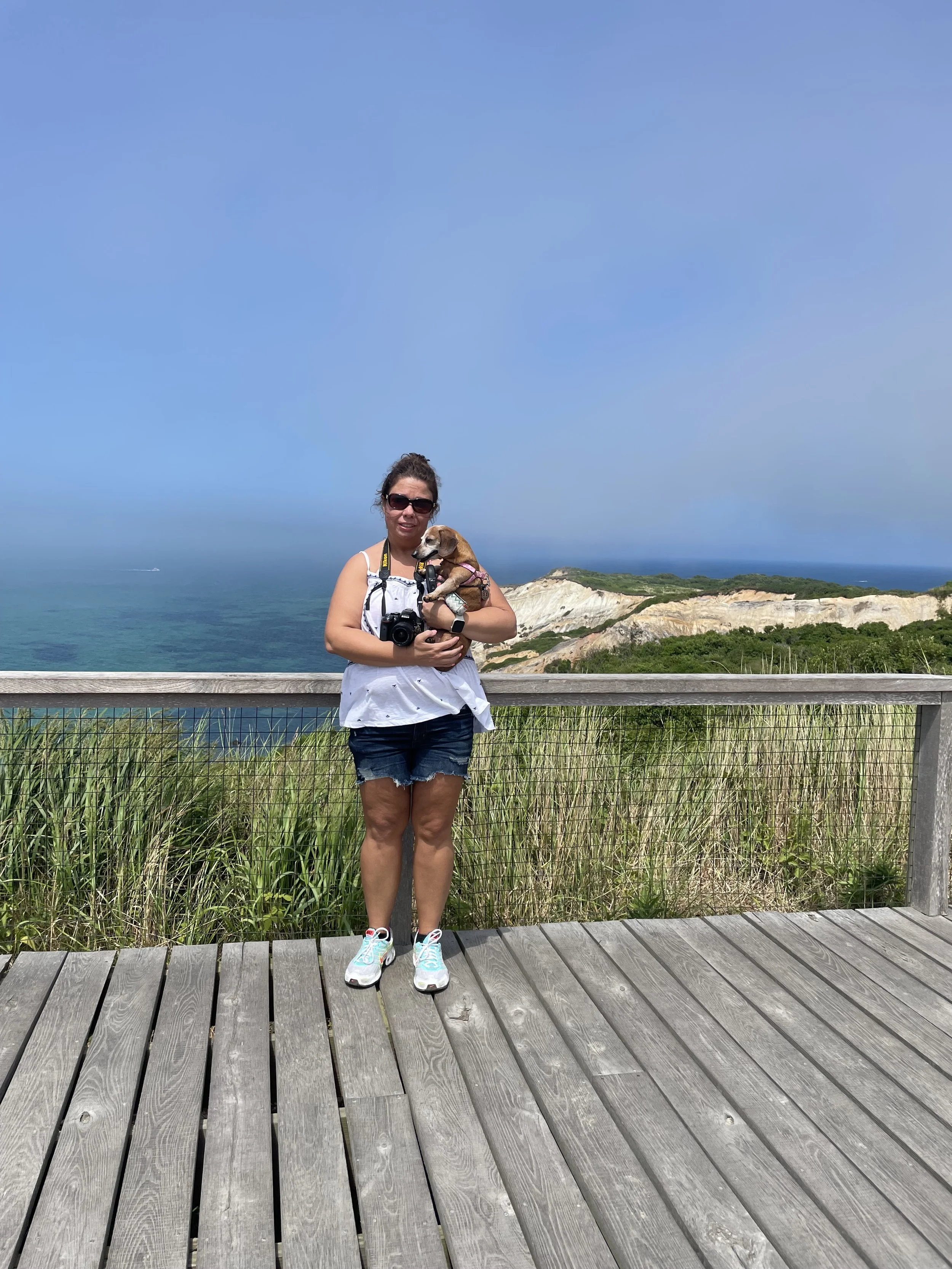 A woman standing on a wooden viewing platform with green foliage and cliffs behind her, holding a small dog and a camera, under a of blue sky and ocean.