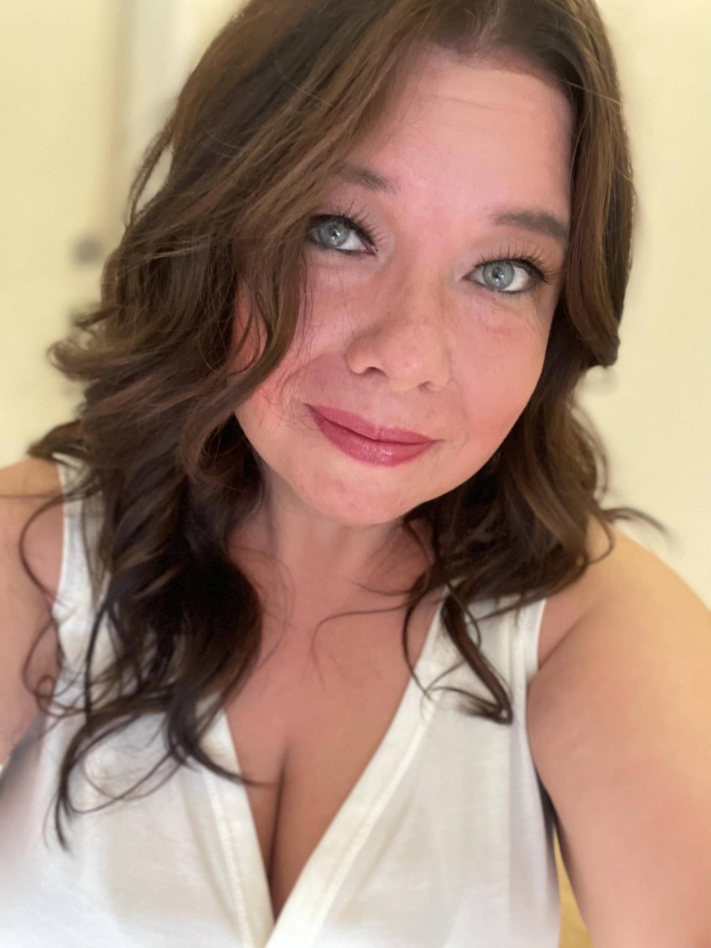 Close-up of a woman with shoulder-length wavy brown hair, blue eyes, and light skin, wearing a white top, smiling softly.
