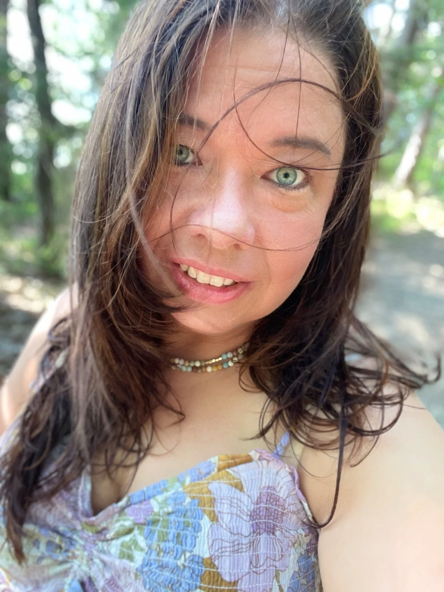 A woman with long, wet brown hair and green eyes is smiling outdoors in a wooded area, wearing a floral top and a beaded necklace.