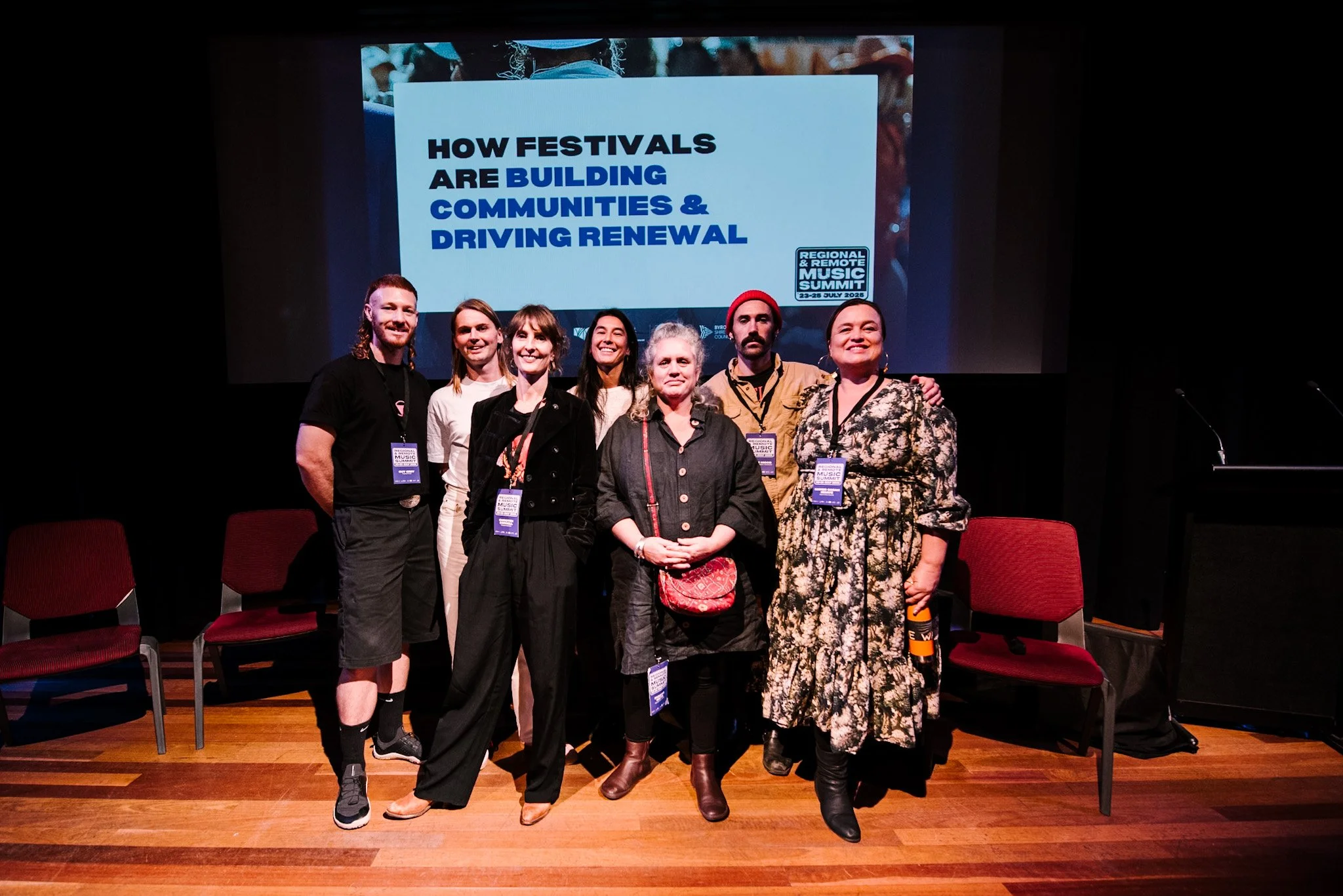 Group of seven people standing on stage in front of a large screen at the Regional & Remote Music Summit, with the text 'How festivals are building communities & driving renewal' displayed. The group is smiling and wearing conference badges.