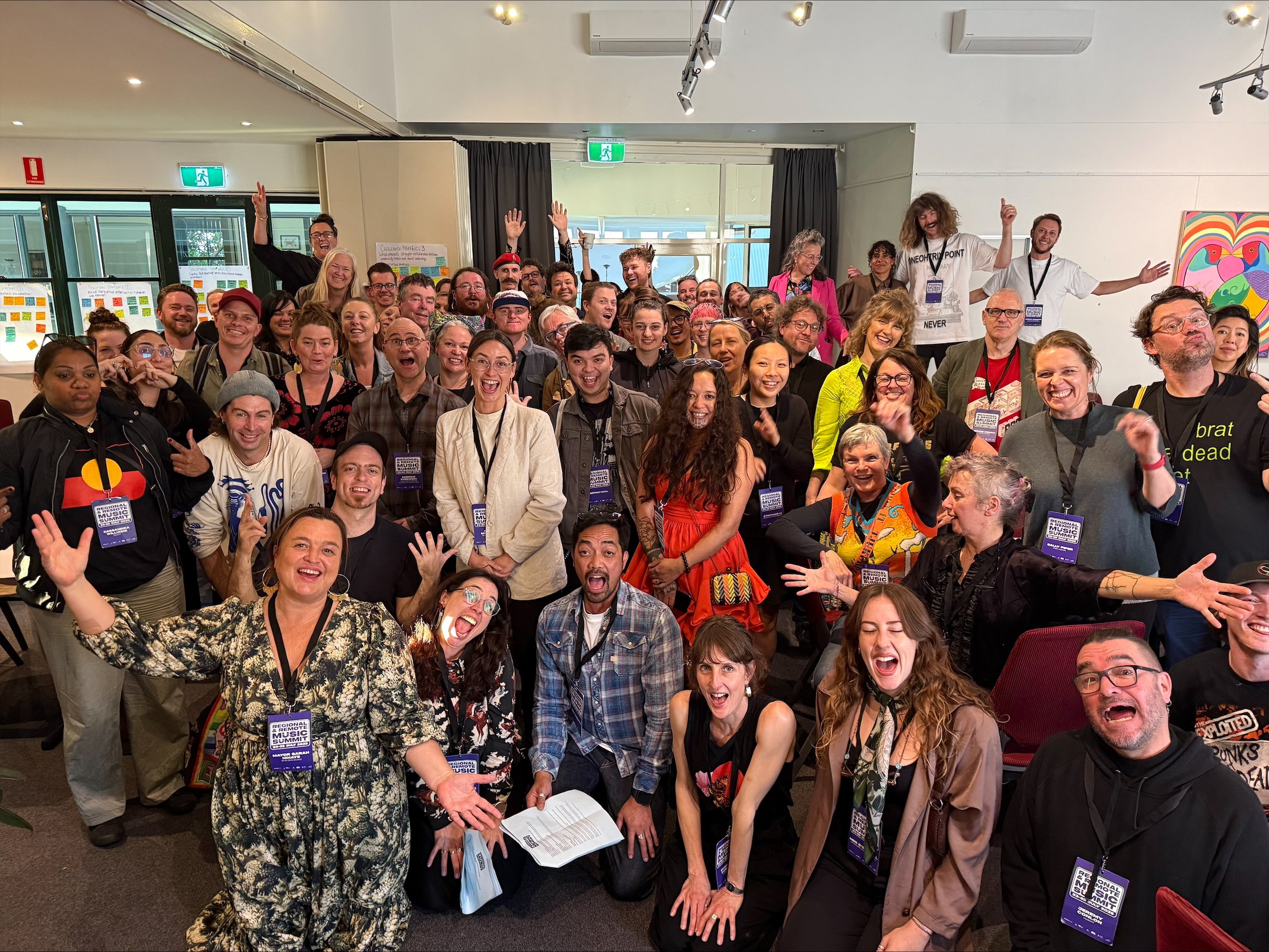 A large group of people gathered indoors, smiling, cheering, and posing for a group photo at a conference or event, with some holding identification badges.
