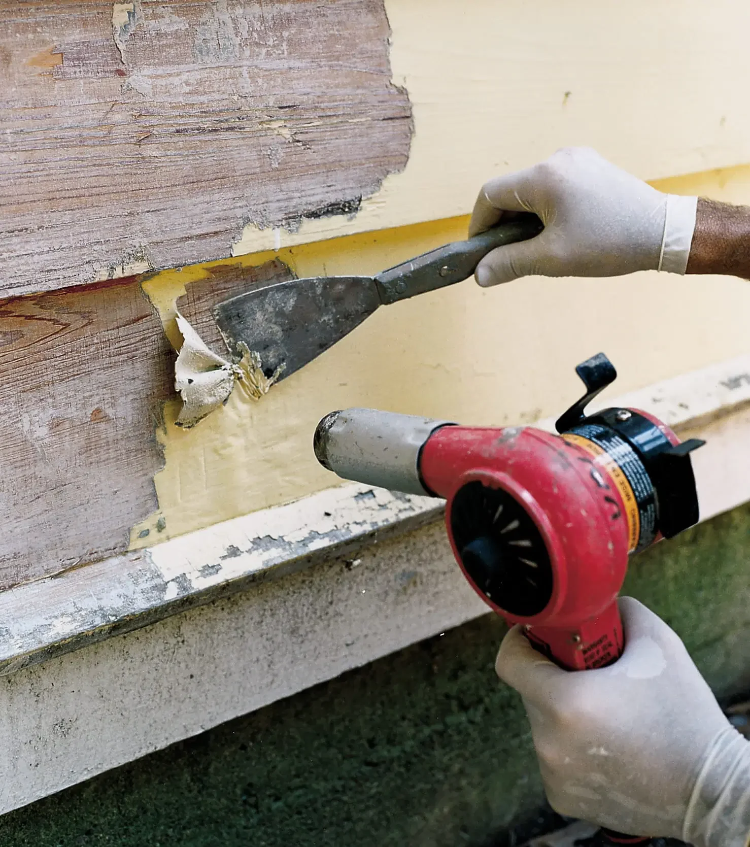 Person wearing gloves using a heat gun on a wooden surface while removing paint or varnish.
