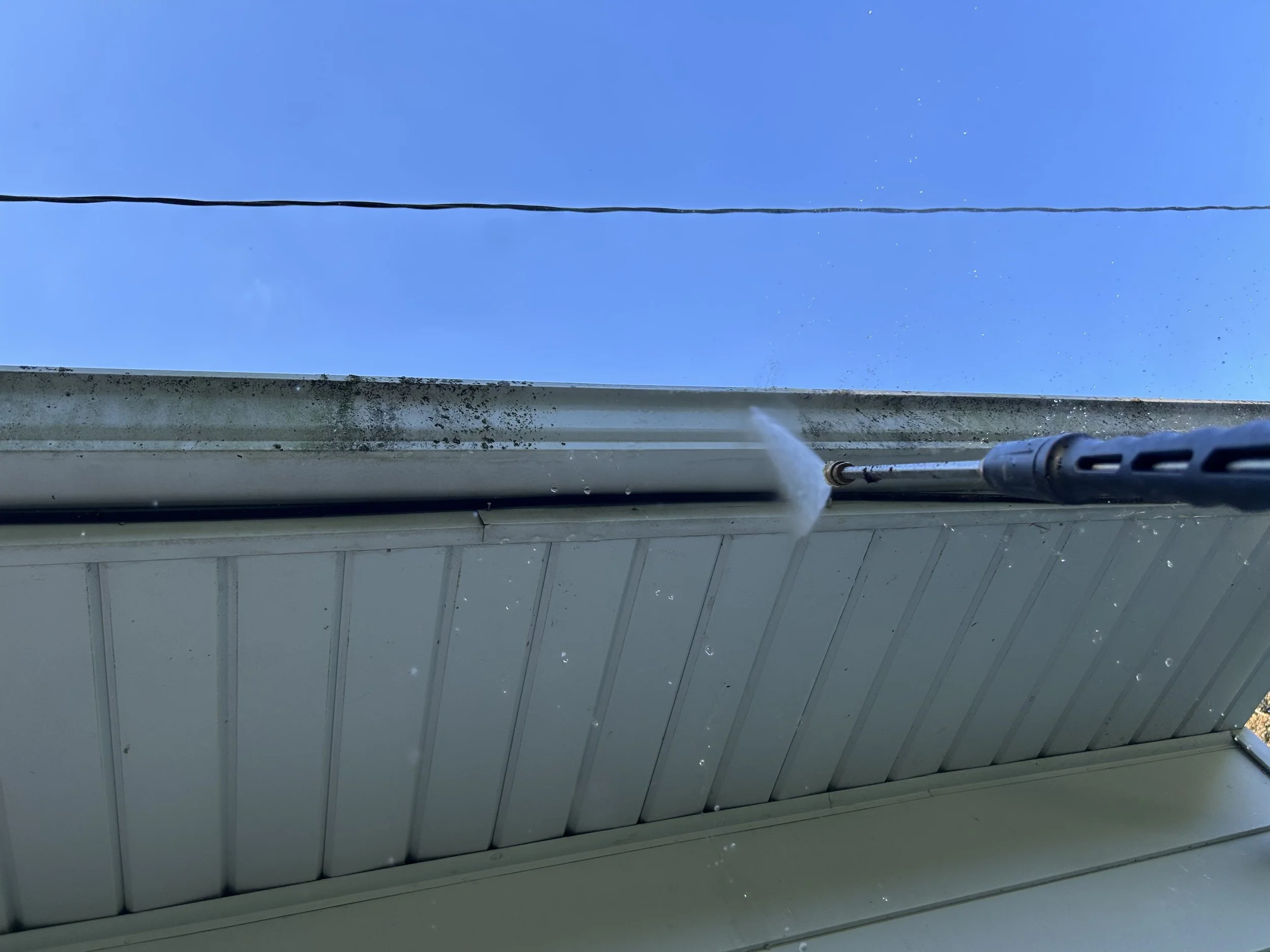 Person using a pressure washer to clean the underside of a roof overhang outdoors with a clear blue sky in the background.
