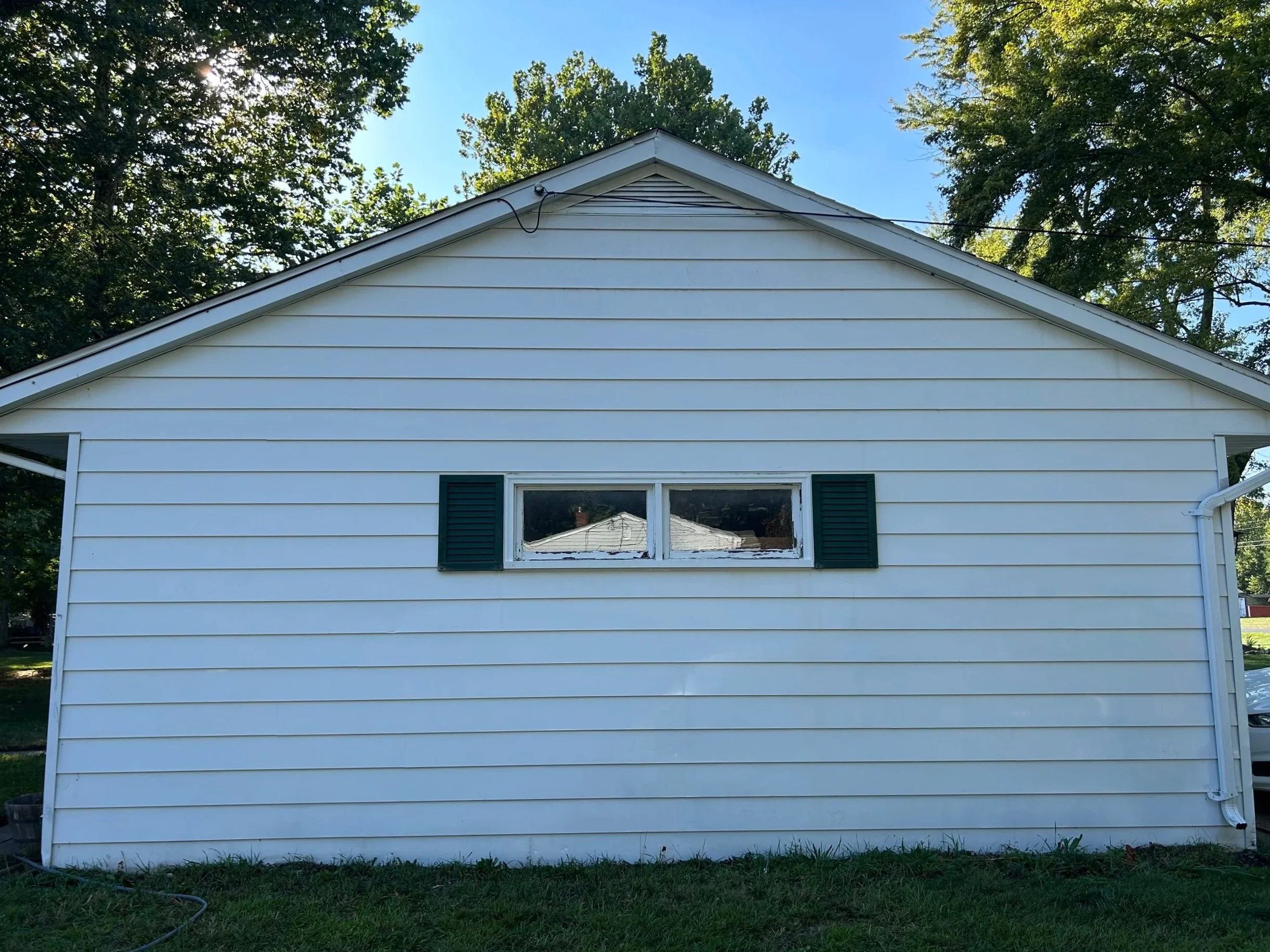 Front of a white house with horizontal siding, a sloped roof, and a narrow rectangular window with dark green shutters.