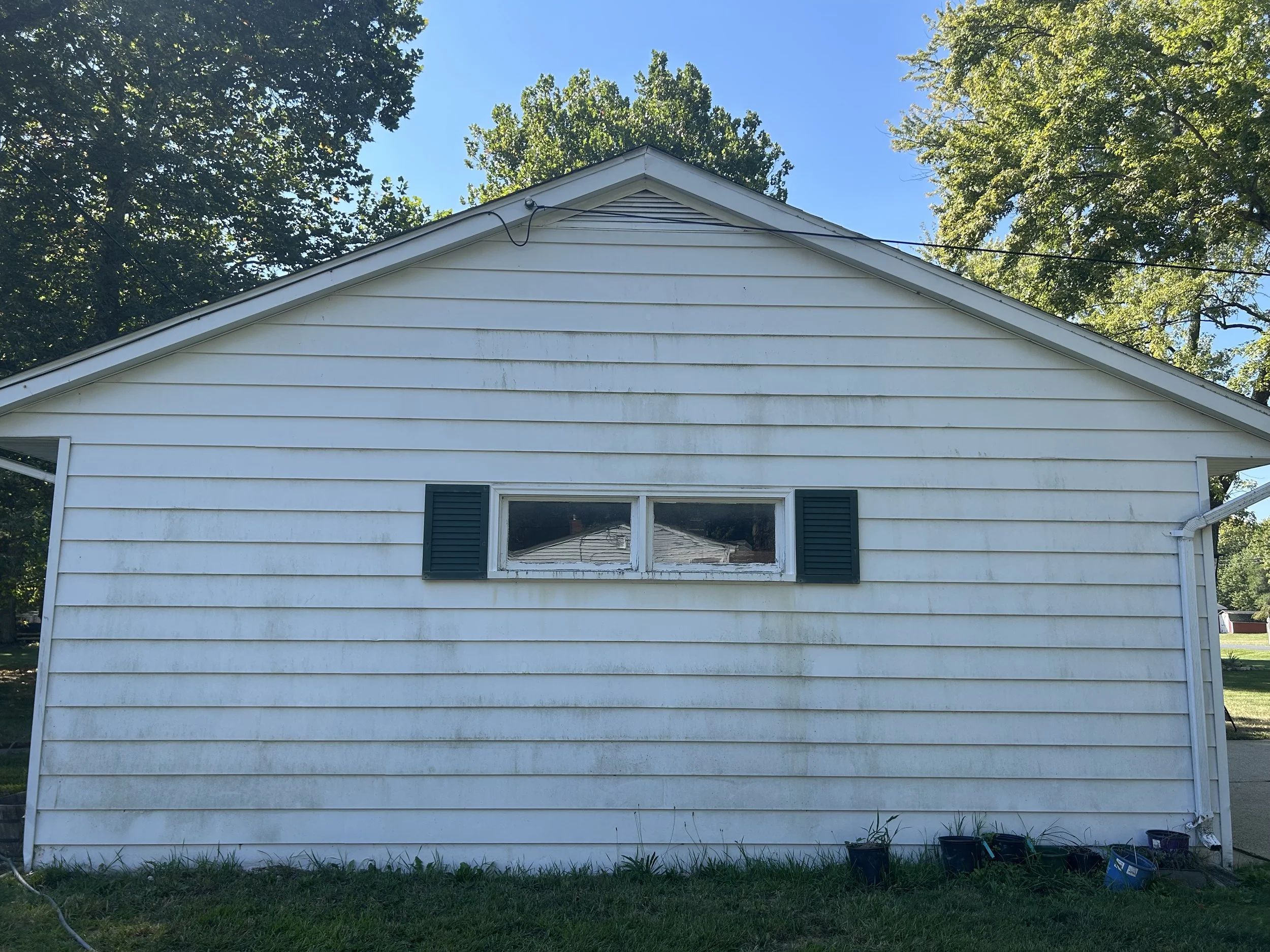Front view of a white house with horizontal siding, a small rectangular window with two panes and dark green shutters, and a roof with trees behind it. There are several planting pots at the bottom right corner.