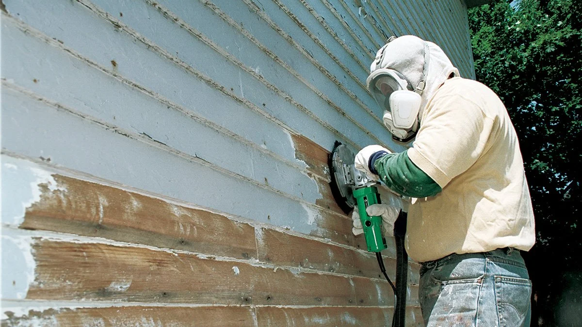 A person wearing a protective mask and gloves is sanding the exterior of a house with wooden siding.