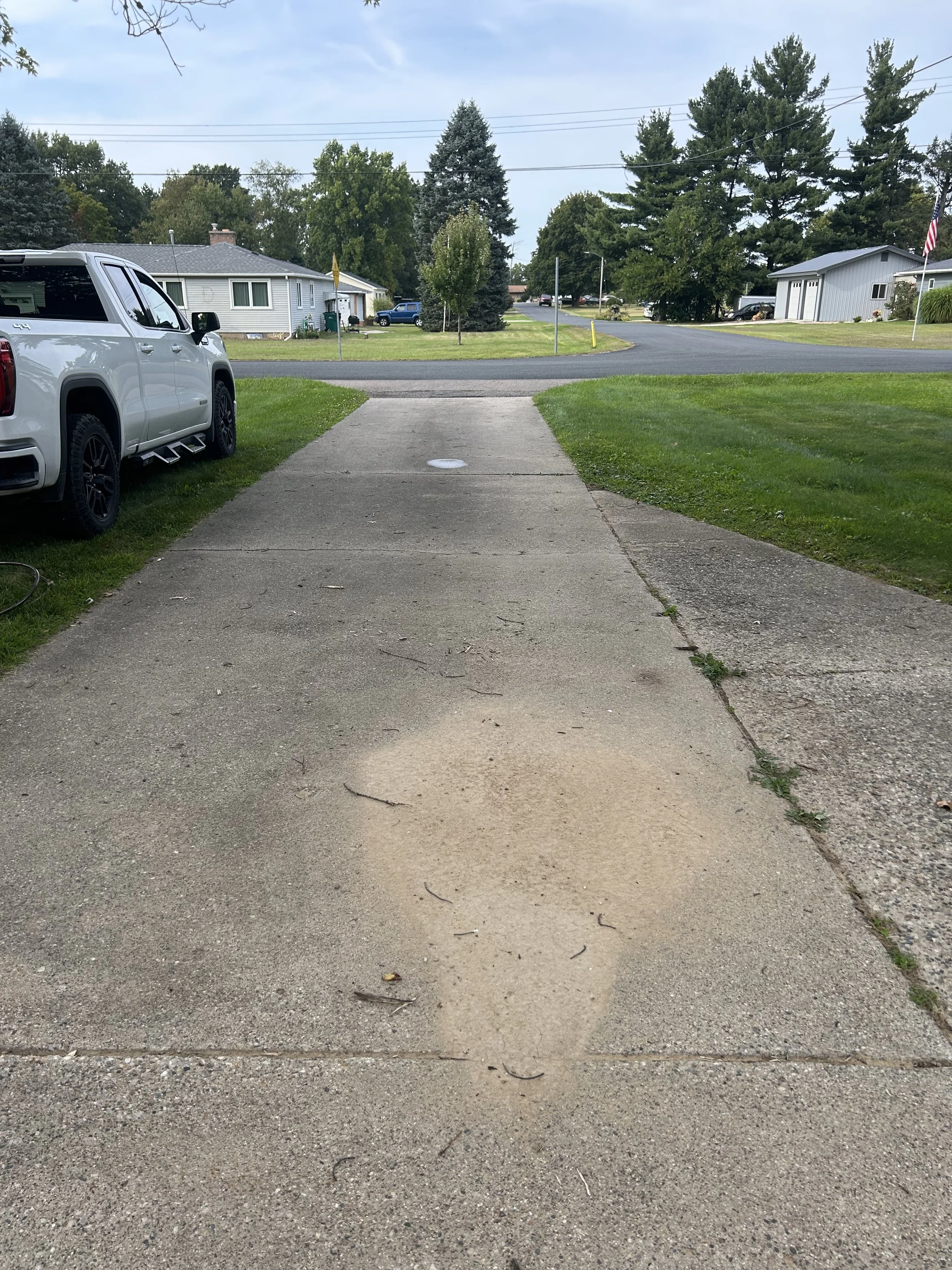 A concrete driveway leading to the street in a suburban neighborhood with green lawns, trees, and houses. A silver pickup truck is parked on the left side.