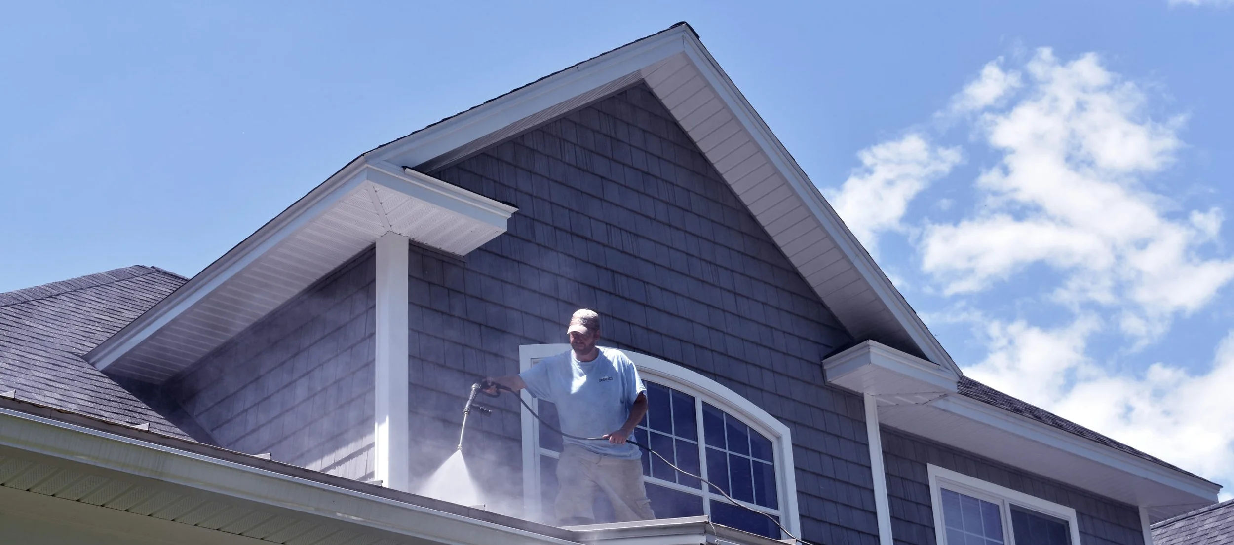 A man power washing the roof of a house with blue shingles and white trim, under a blue sky with some clouds.