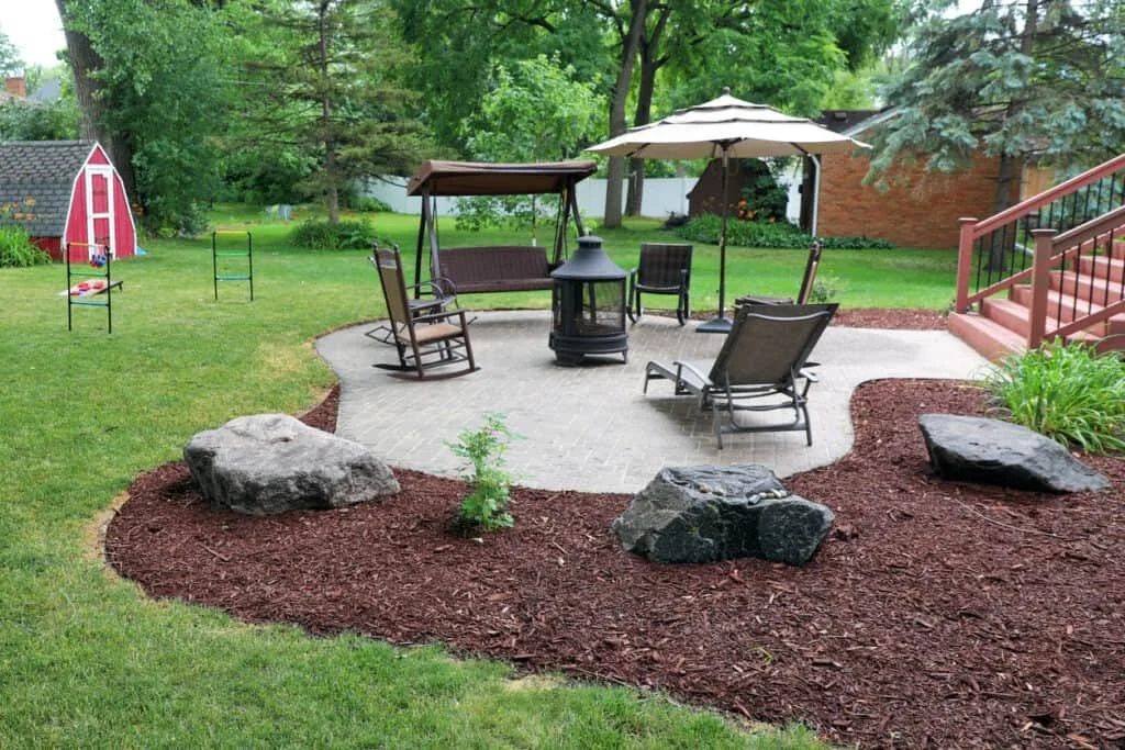 Backyard with a patio area featuring outdoor chairs, a table with an umbrella, and a fireplace. Surrounding the patio are large rocks, mulch beds, a slide and climbing equipment on the grass, and a small red shed. There are trees and a house in the background.
