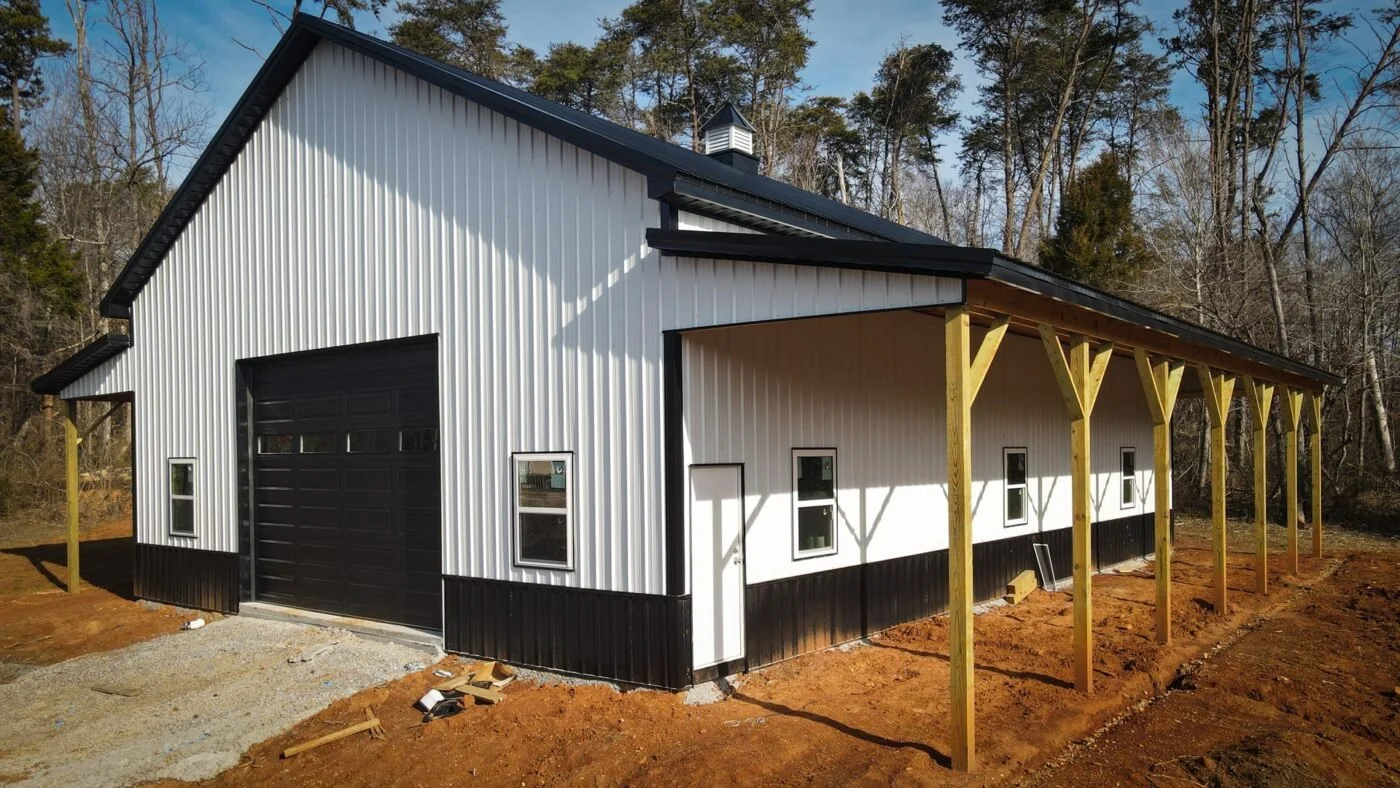 Newly constructed barn with white metal siding, black accents, and a large garage door, surrounded by trees and a clear sky.
