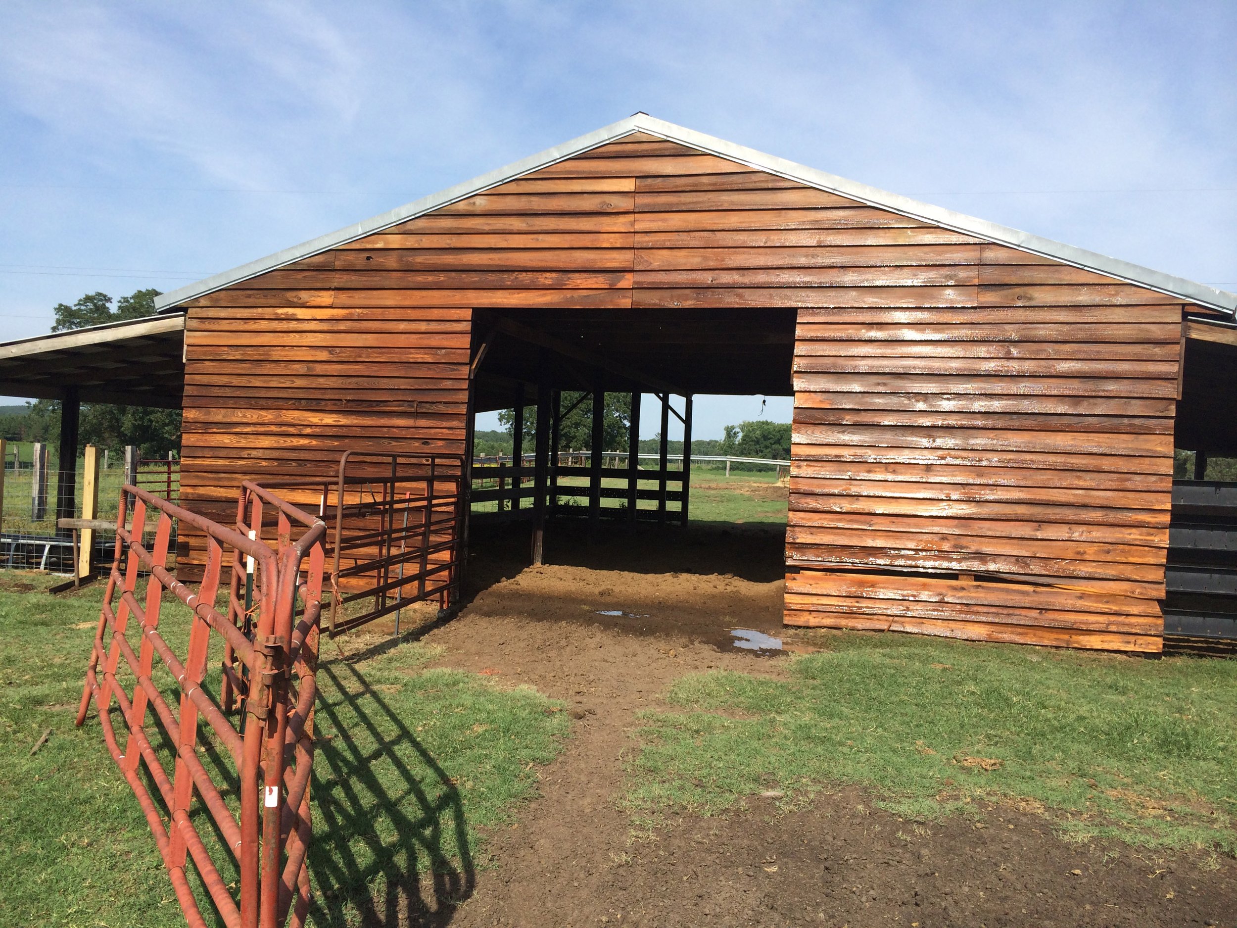 A wooden barn with an open entrance, surrounded by a metal red gate and green grass, in a rural setting under a blue sky.