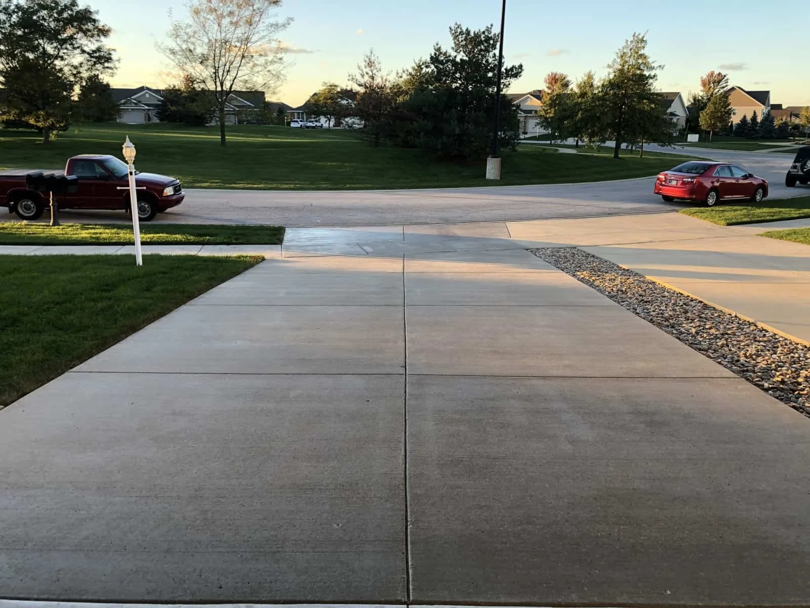 View from a driveway showing a sidewalk, a street with parked cars, and green lawns with trees in a suburban neighborhood during daytime.