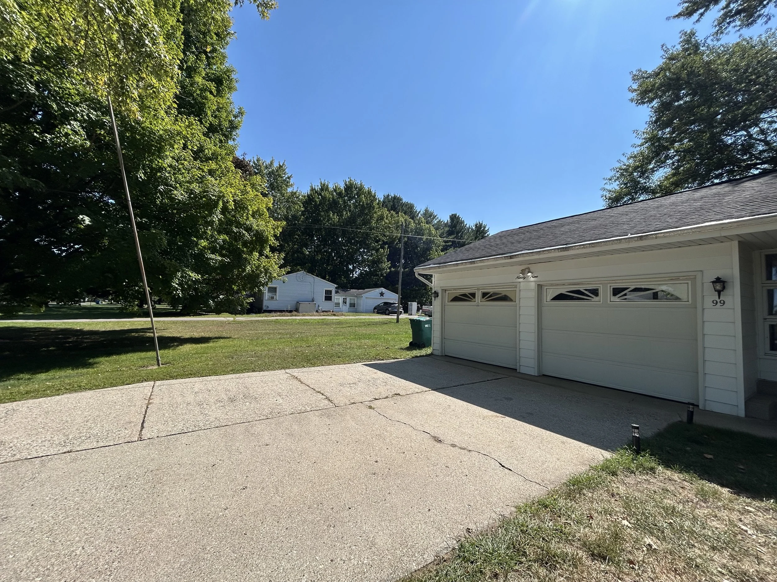 View of a suburban house's driveway and garage, with a large tree on the left, a clear blue sky, and neighboring houses in the background.
