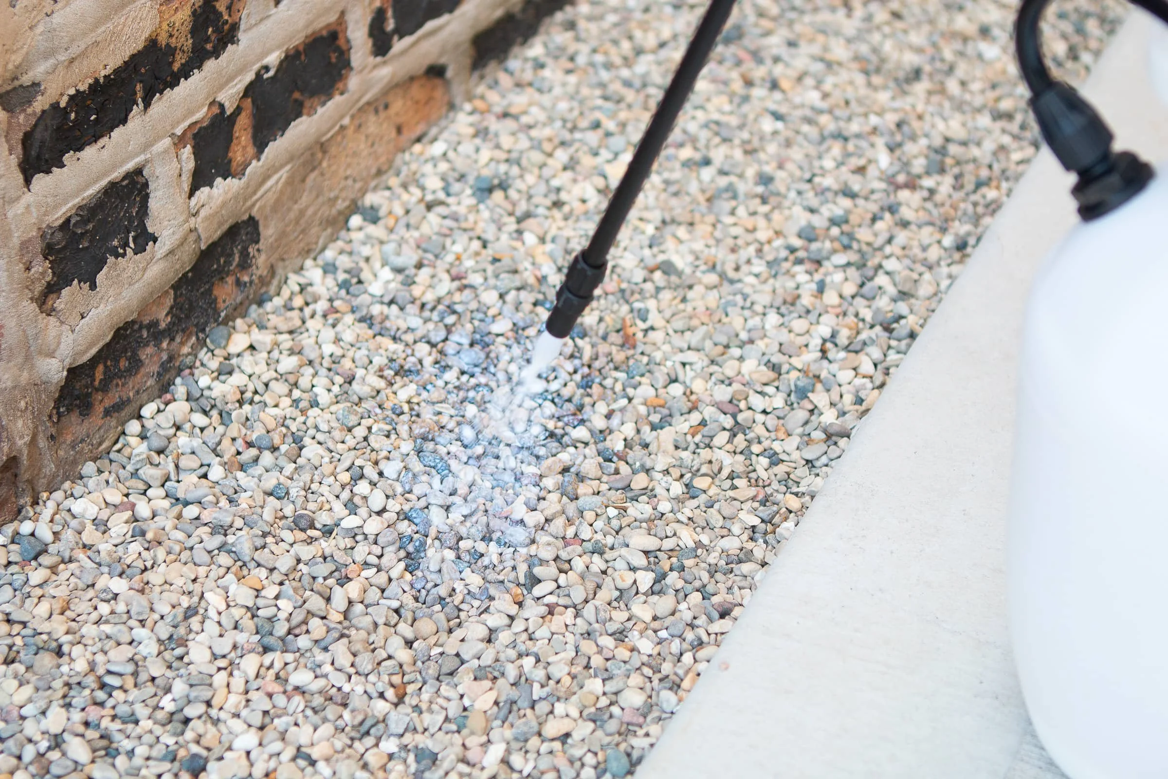 Close-up of a pressure washer spraying water on small gravel near a brick wall and concrete sidewalk.