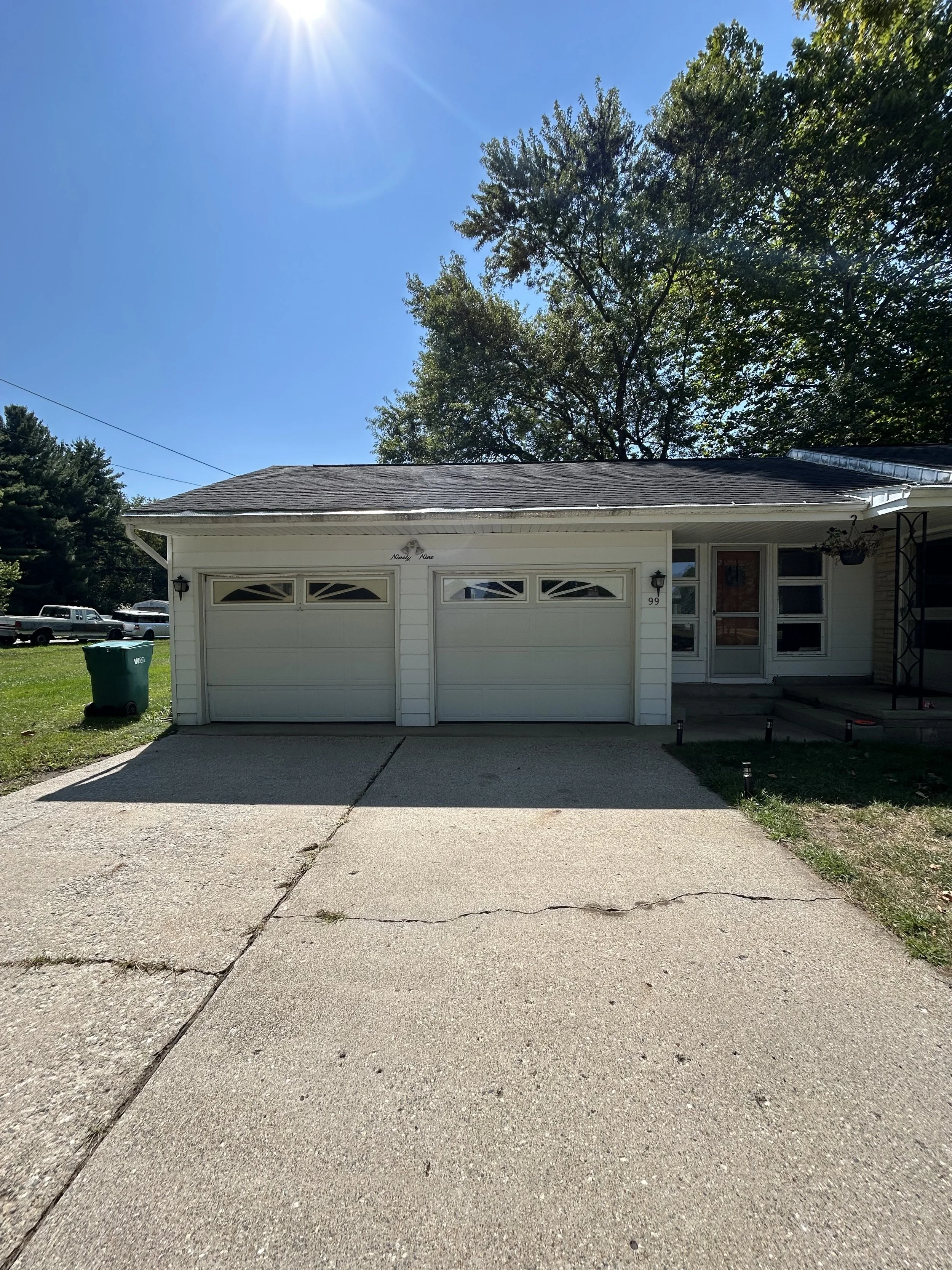 Front of a white house with a double garage, a concrete driveway, and a small porch with stairs. There are trees behind the house and a clear blue sky with the sun shining.