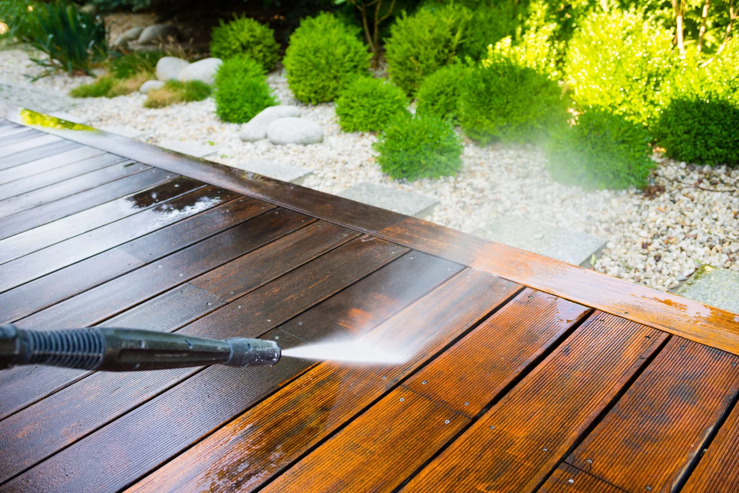 A person is pressure washing a wooden deck outdoors, with a garden featuring green bushes and white rocks in the background.