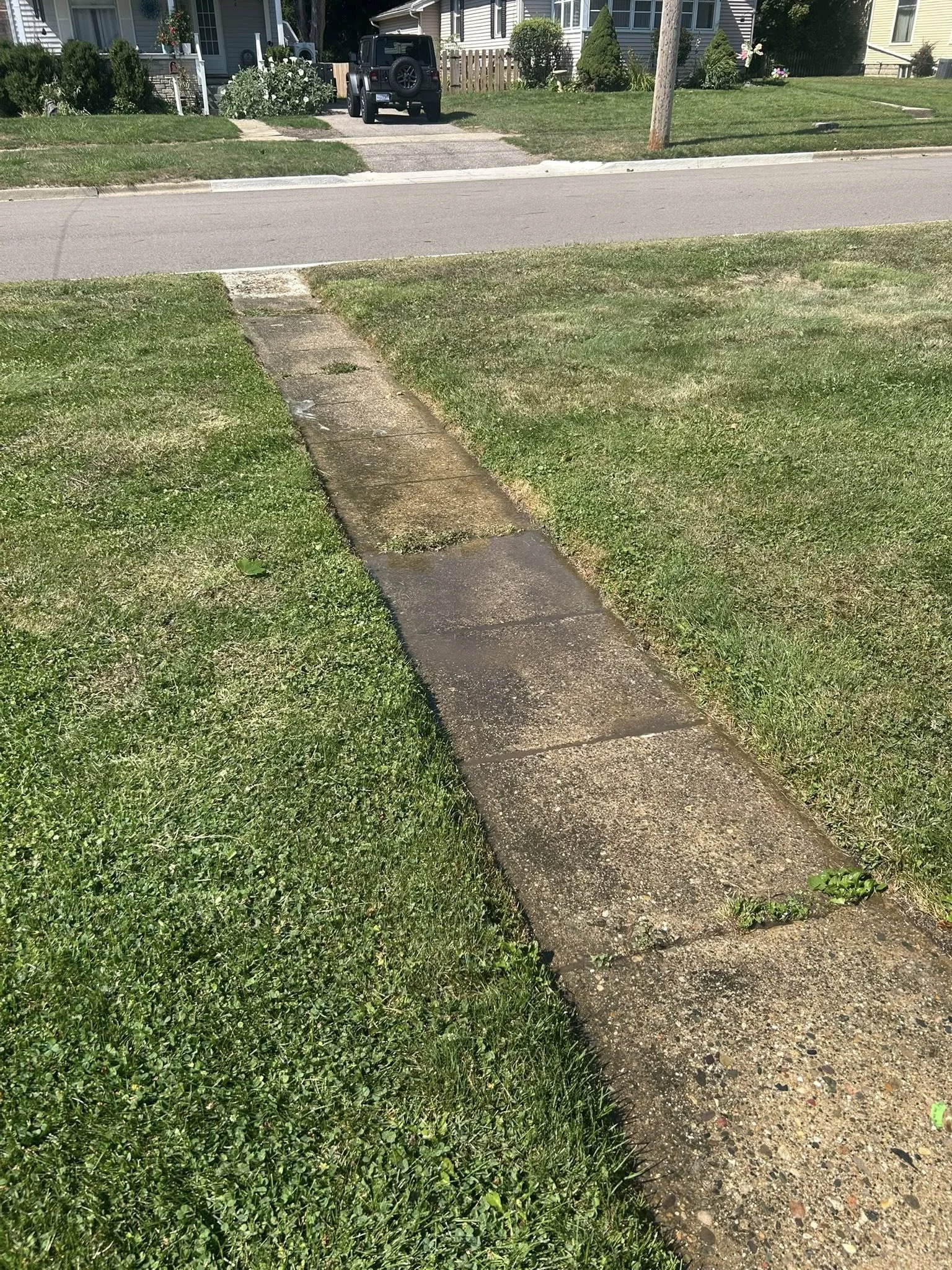 Concrete sidewalk running through green grass in a residential neighborhood with houses, trees, and a parked vehicle in the background. before cleaning