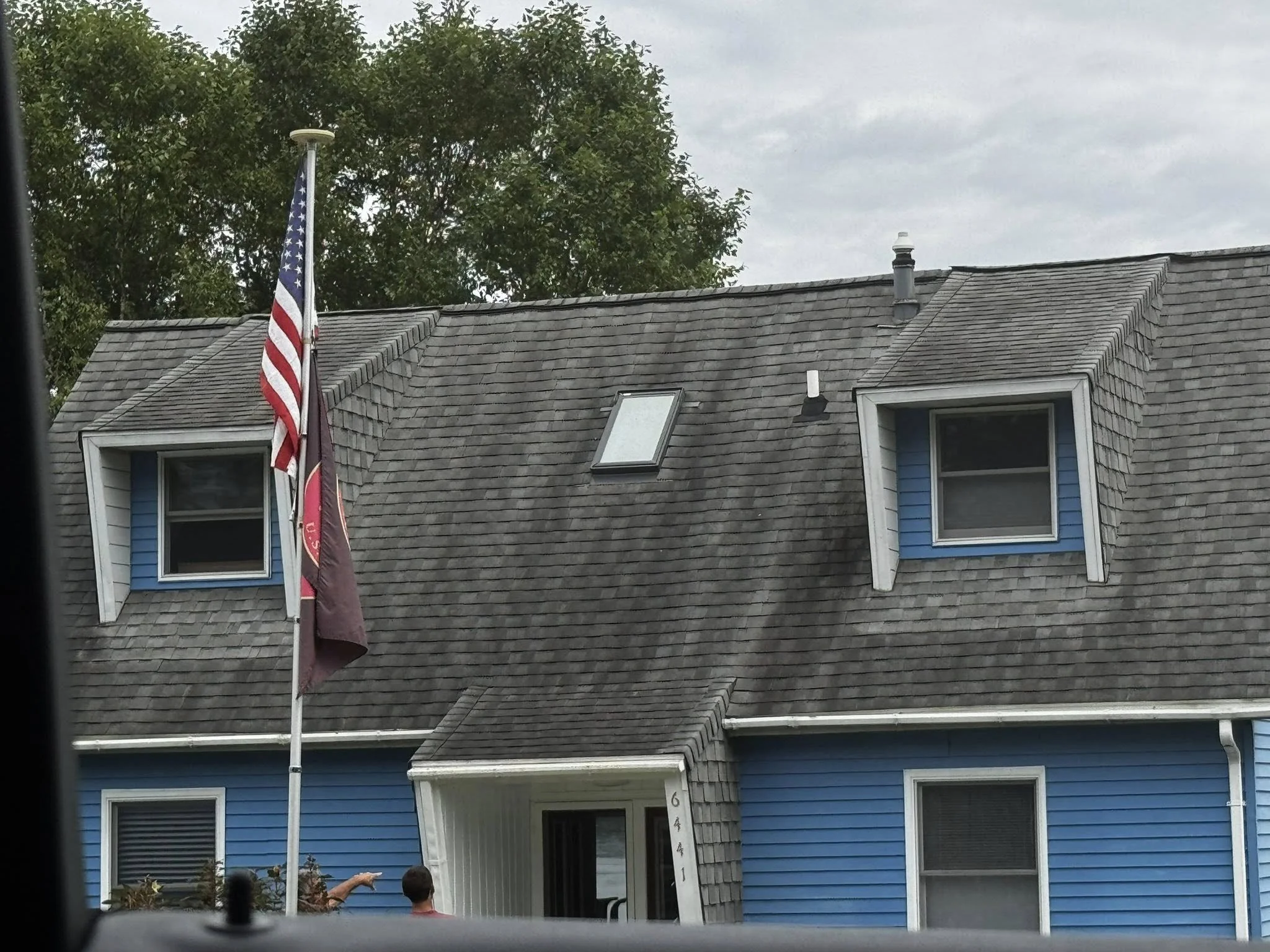 A blue house with a gray shingle roof, two dormer windows, and a chimney, with an American flag flying in front.