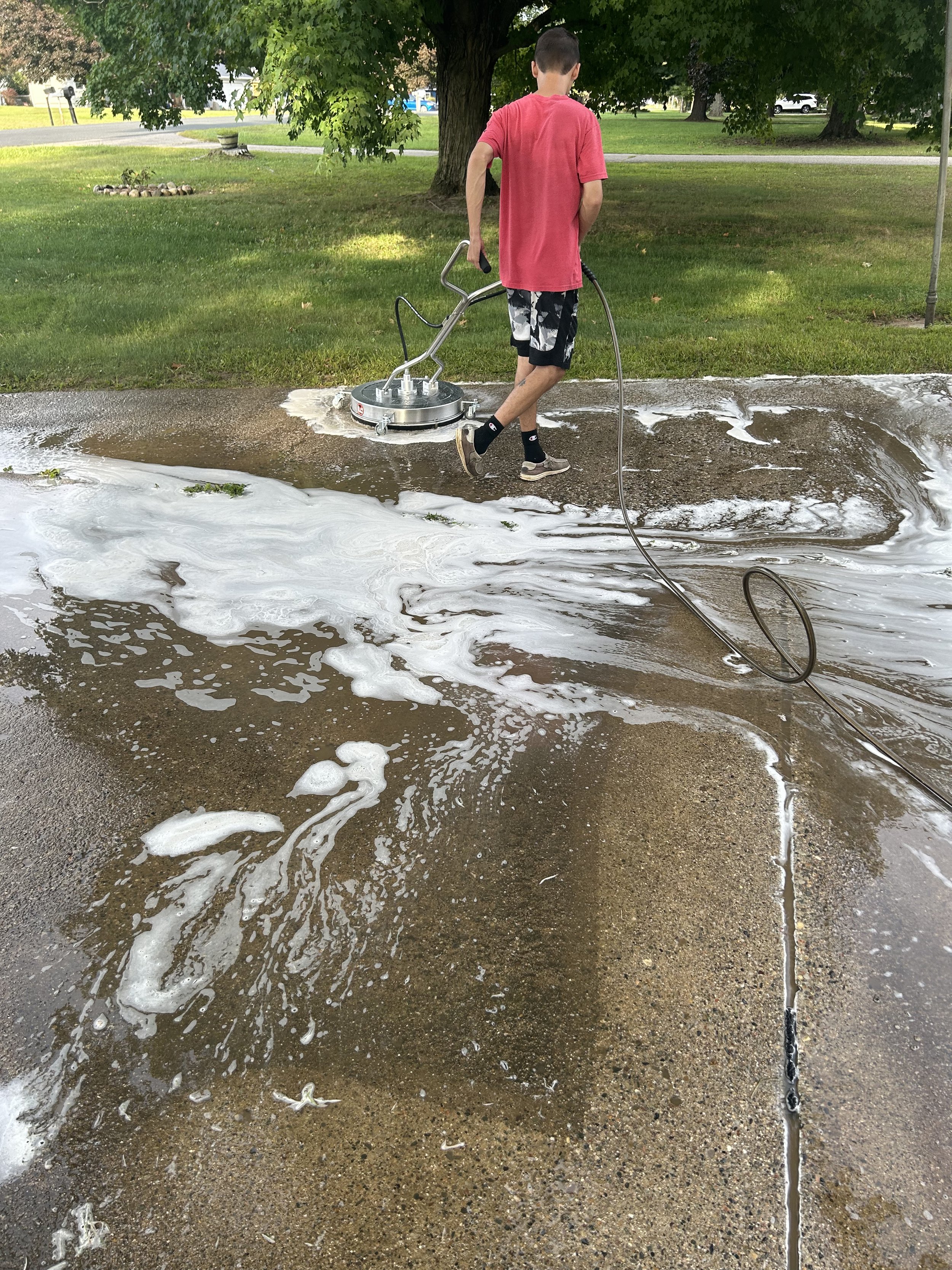 Person cleaning a concrete driveway with a pressure washer, creating soap and foam on the surface.