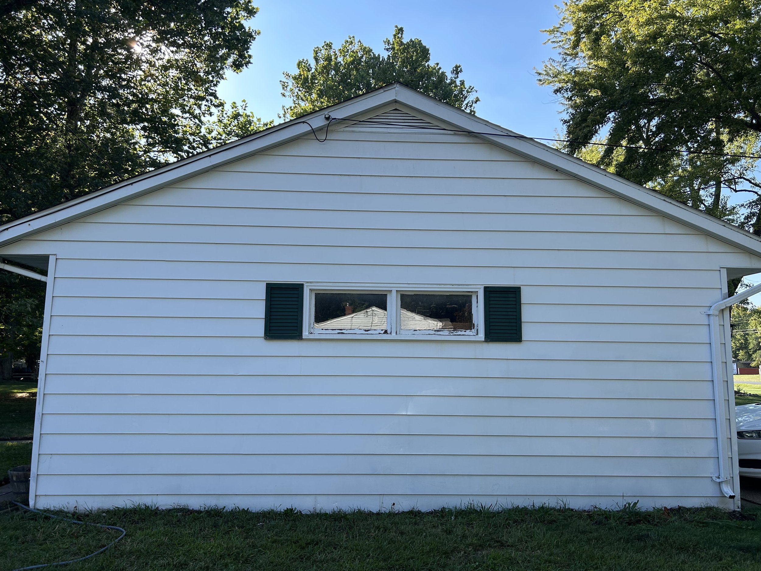 The side view of a white house with horizontal siding, green shutters on a small rectangular window, and a sloped roof, with trees in the background and a lawn in the foreground.