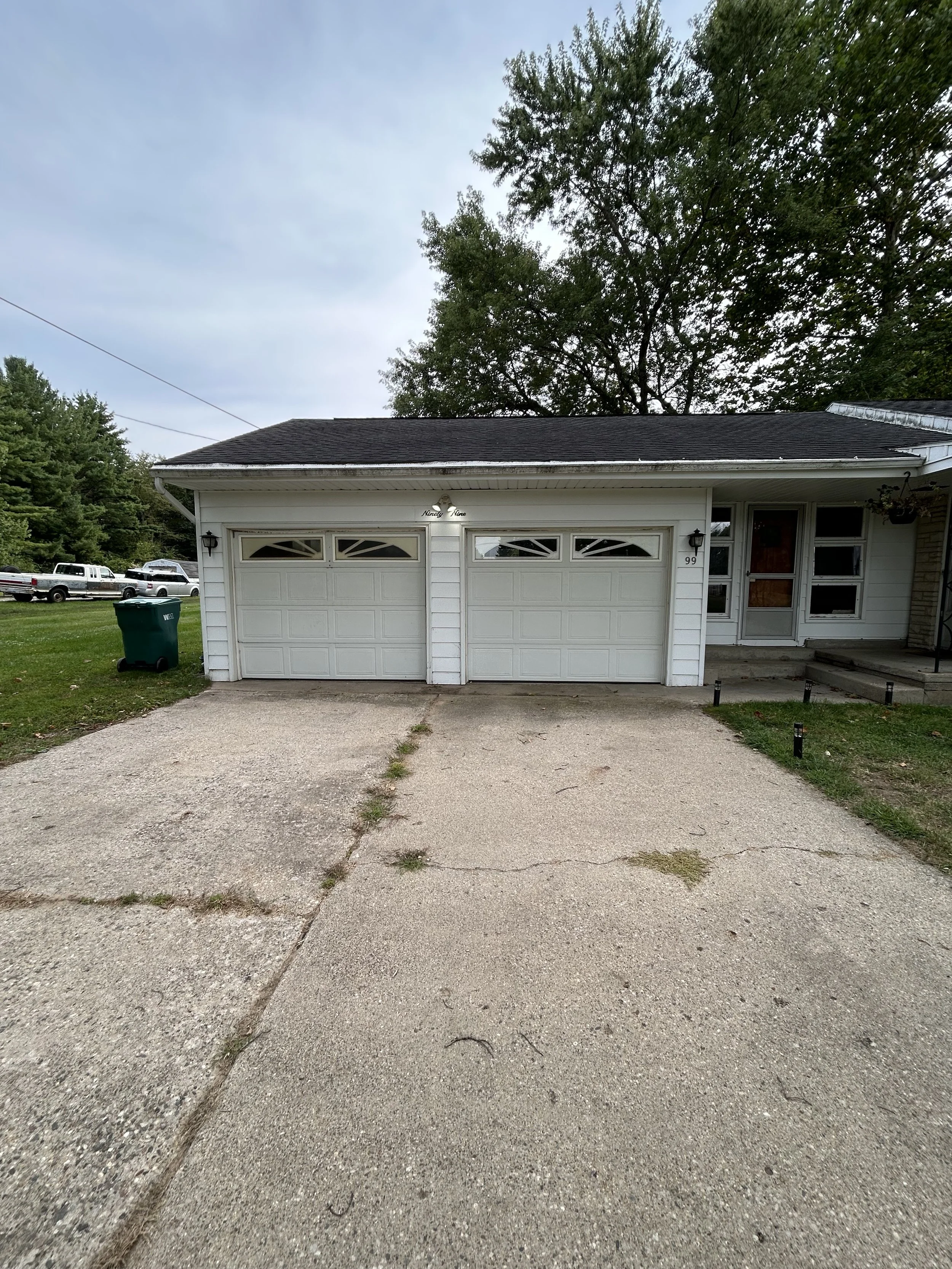 Front view of a house with a double garage and a driveway in the foreground, surrounded by trees and a lawn on the left side, with a trash bin on the side of the driveway, under a cloudy sky.