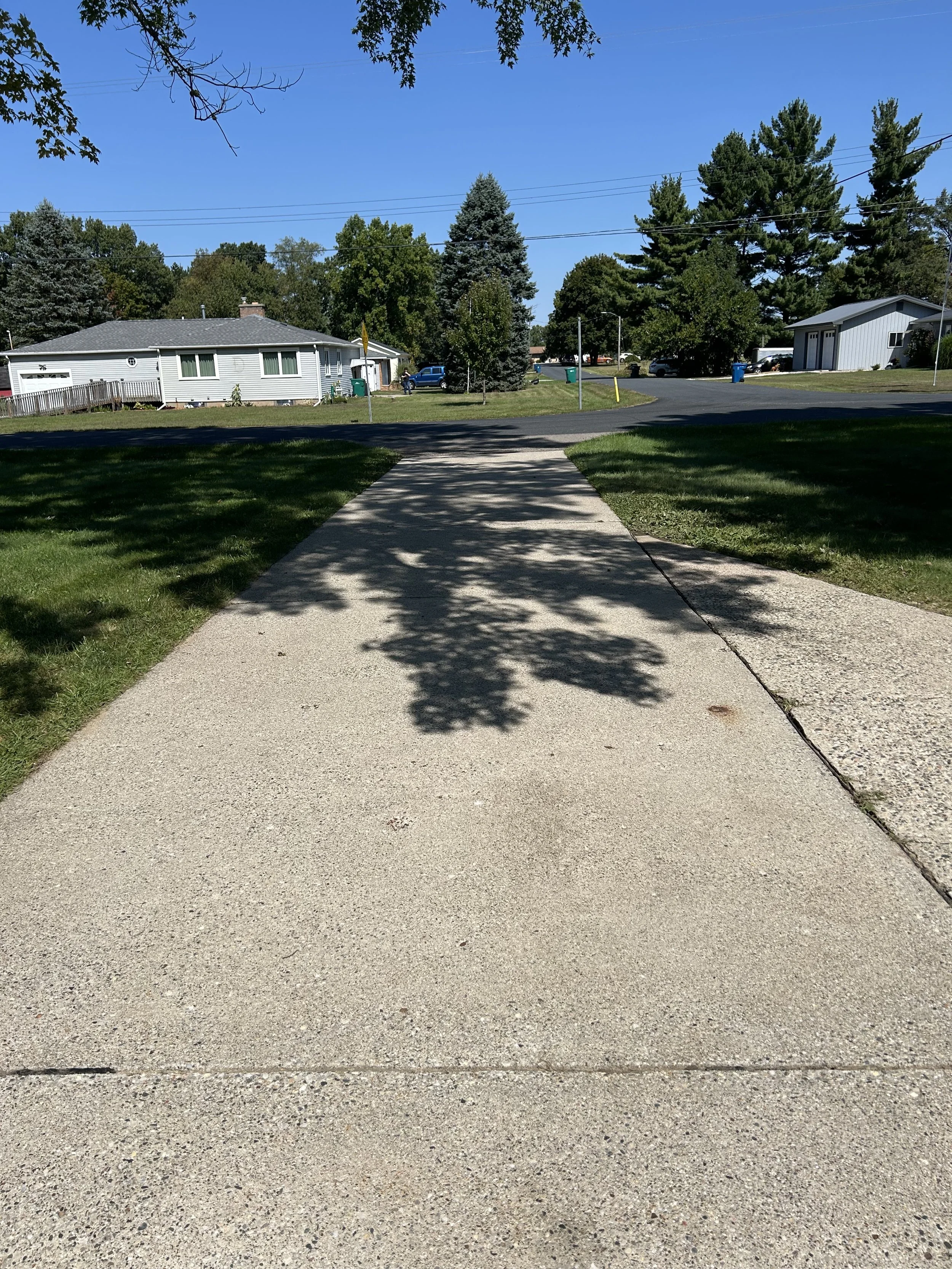 A sidewalk leading away from the camera with shadows of tree branches on it. There are green lawns on either side and suburban houses in the background under a clear blue sky.