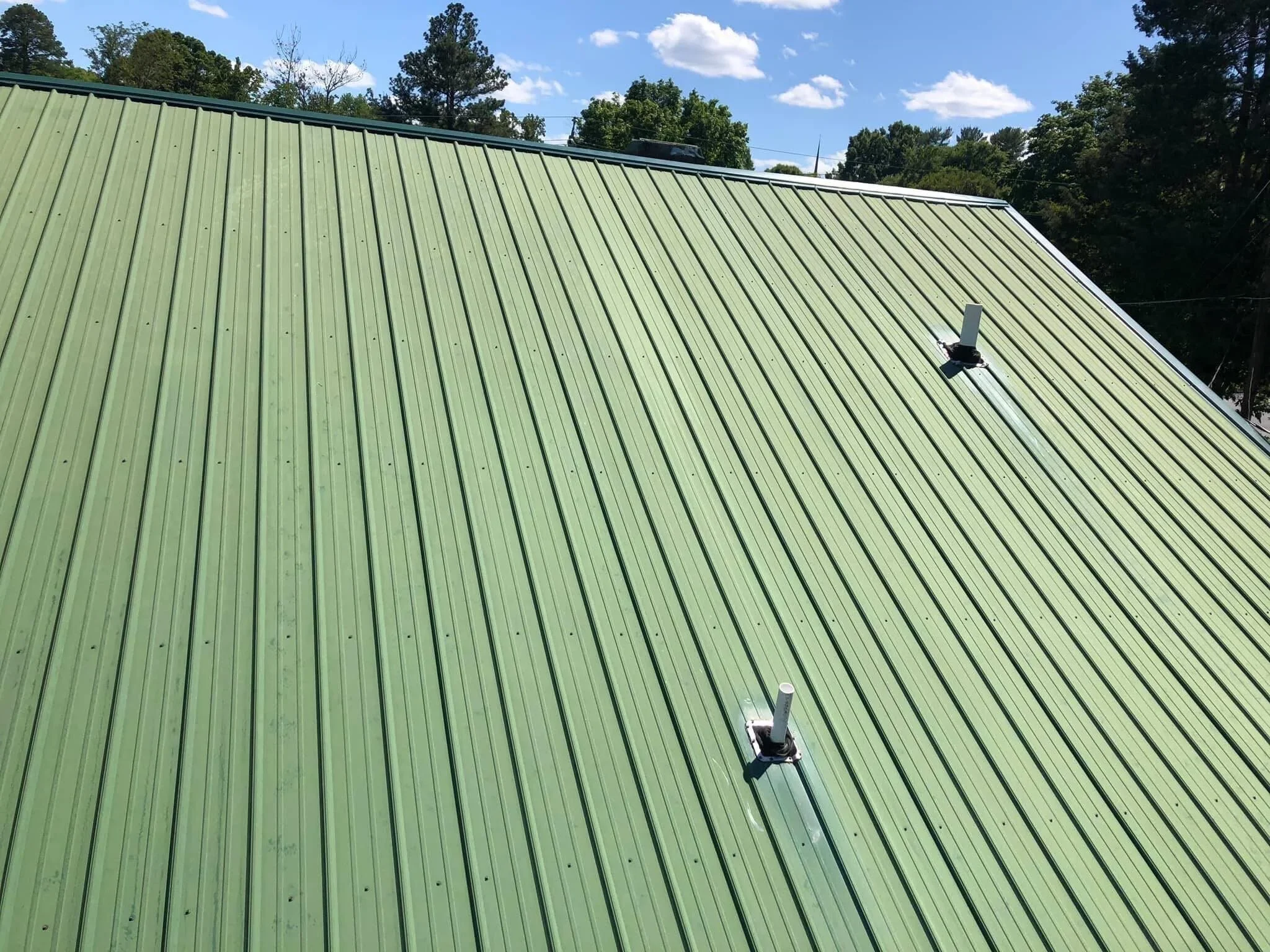 Green metal roof with two vent pipes and a background of trees and blue sky with clouds.