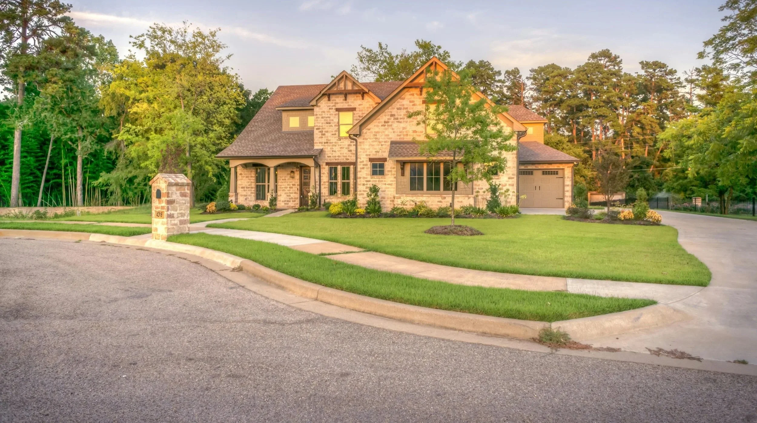 A large, two-story brick house with a gable roof, several windows, and a garage, surrounded by a well-maintained lawn, trees, and shrubs, in a suburban neighborhood during daylight.