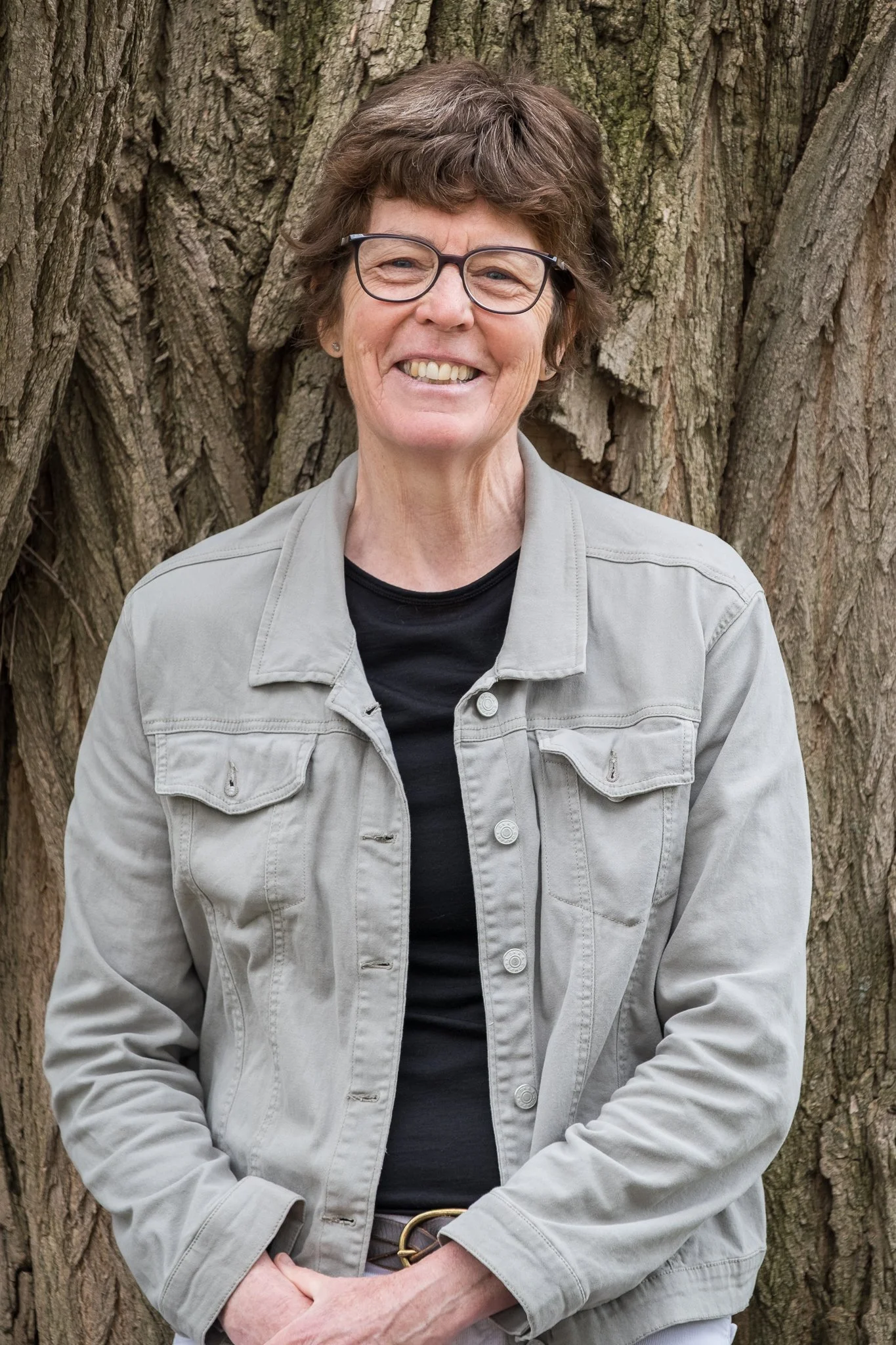 A smiling woman with short brown hair, glasses, and wearing a light grey jacket over a black top, standing outdoors in front of a large tree trunk.