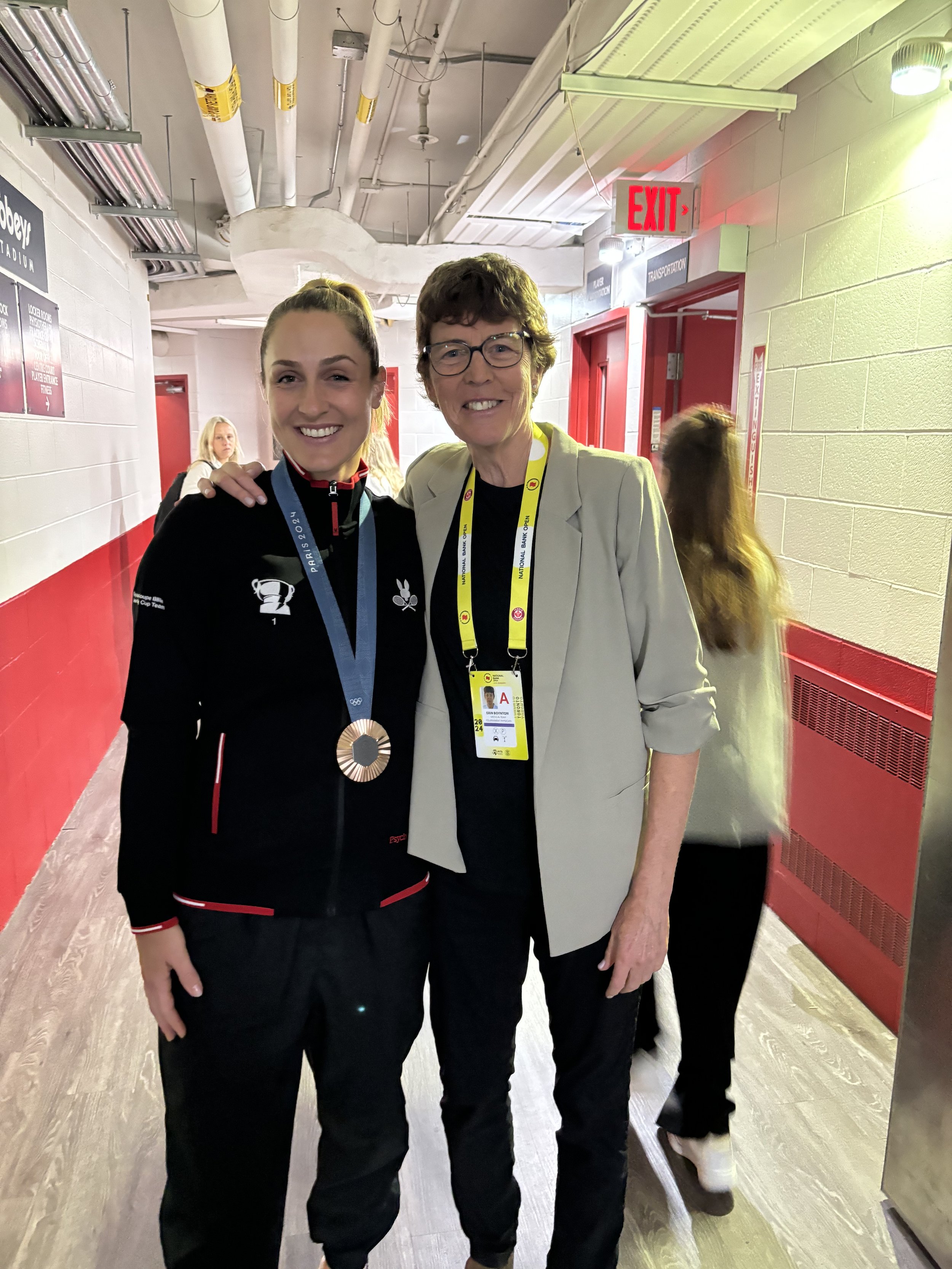 Two women standing in a hallway, smiling at the camera. One woman is wearing a black jacket with a medal around her neck, and the other is dressed in a beige blazer with a yellow lanyard and ID badge.