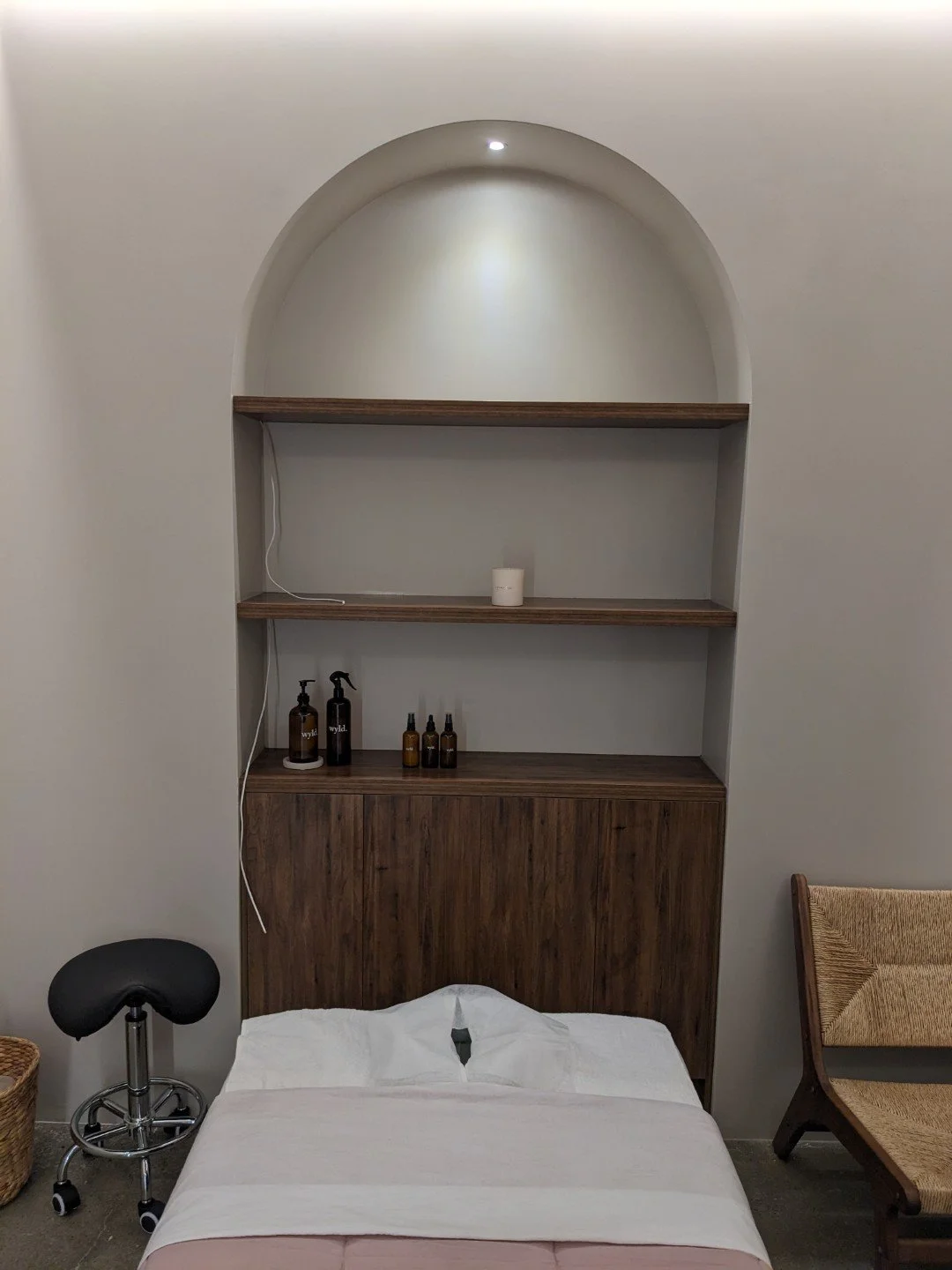 A massage room with a built-in curved wall shelving, cabinets, containing a candle and small bottles, with a bed in front, a black stool on the left, and a woven chair on the right.