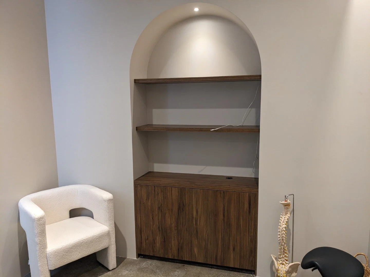 An empty built-in wooden shelves with cabinets in a massage room, a white textured chair to the left, and a human skeleton model and a black chair to the right.