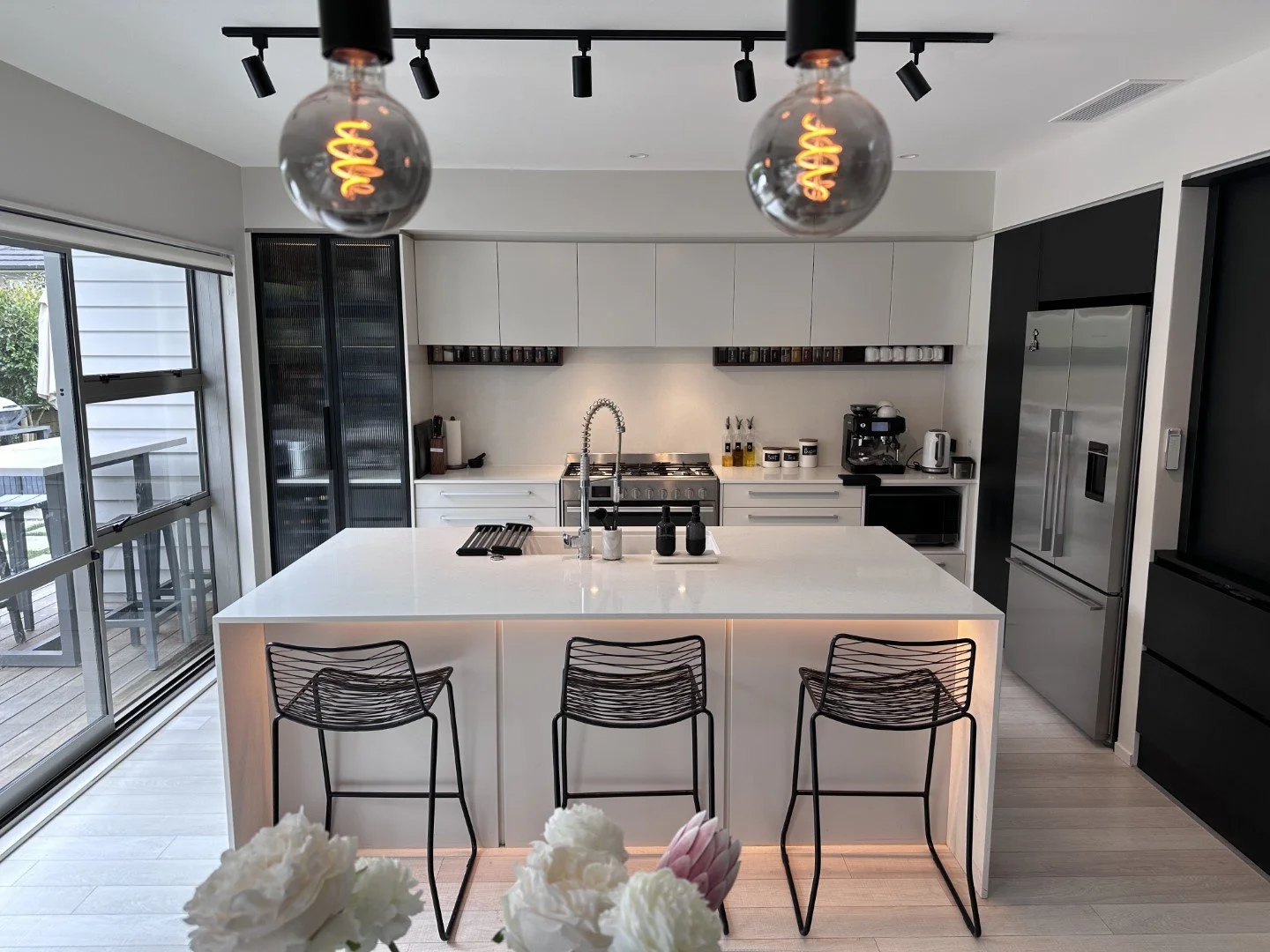 Modern kitchen with a large white island, black wire chairs, and stainless steel appliances, including a refrigerator, oven, and coffee maker, with track lighting and large sliding glass doors leading to an outdoor deck.
