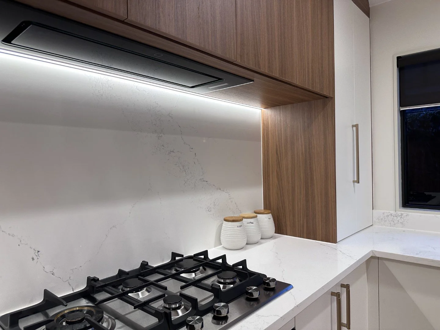 Part of a modern kitchen with white marble countertop, three ceramic jars with wooden lids, black gas hobs, wood and white cabinets, and a window with dark blinds.
