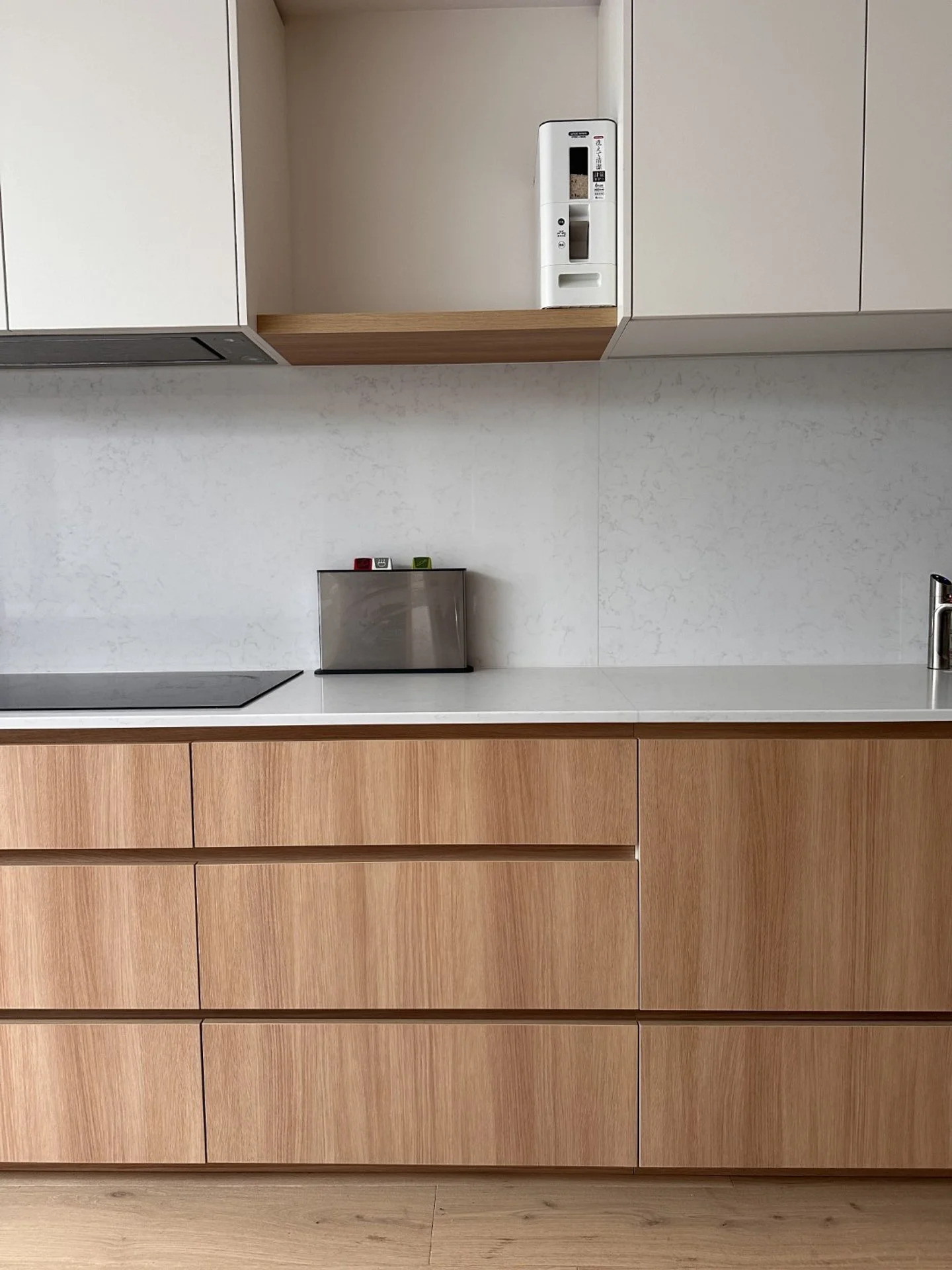 Part of larger kitchen with wooden drawers, a white countertop, and upper cabinets. A small silver appliance with red and green lights sits on the counter. A white wall with subtle textured pattern is in the background.