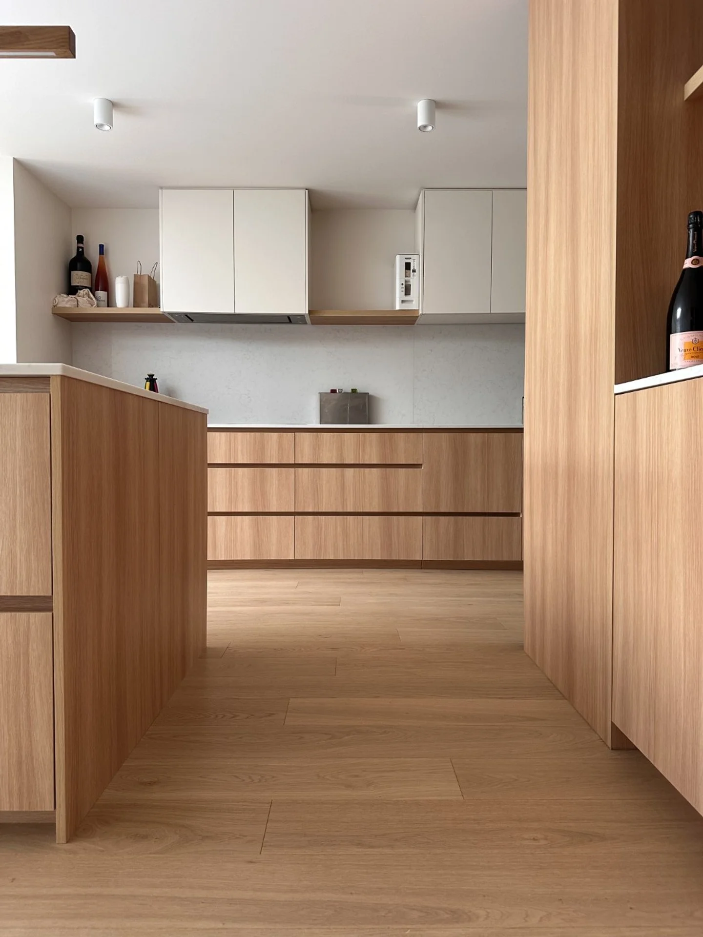 Modern kitchen with wooden cabinetry, white upper cabinets, and hardwood flooring. Items on the counter include bottles and paper bags.