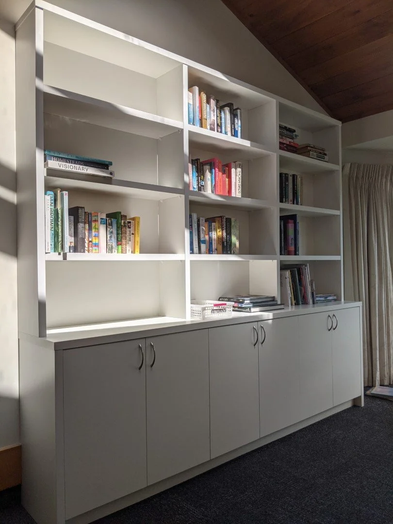 White bookshelf with books and a lower cabinet in a room with beige walls, a dark carpet, and a window with curtains.