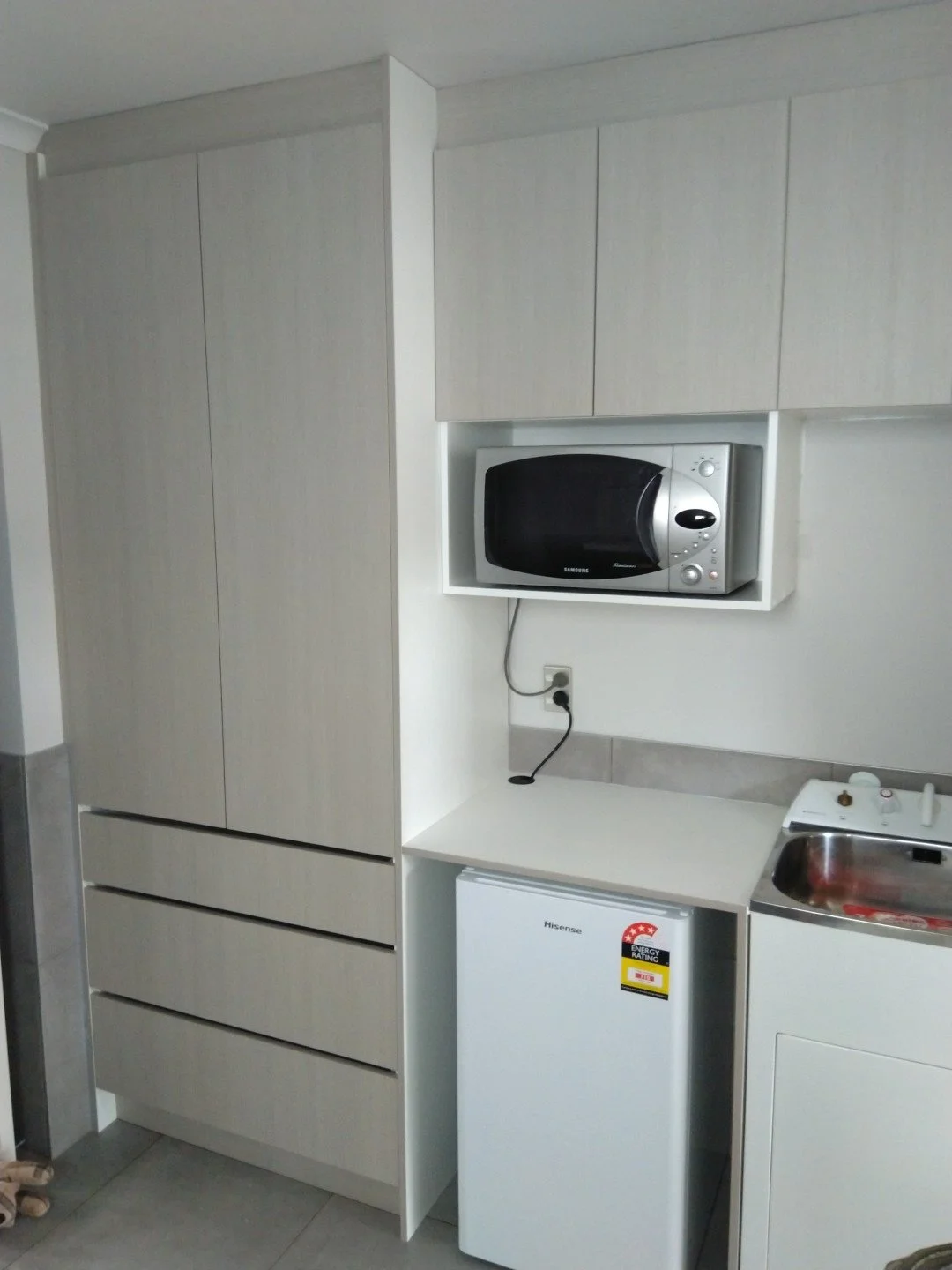 Kitchen corner with mounted microwave oven, small refrigerator, sink, and cabinetry.