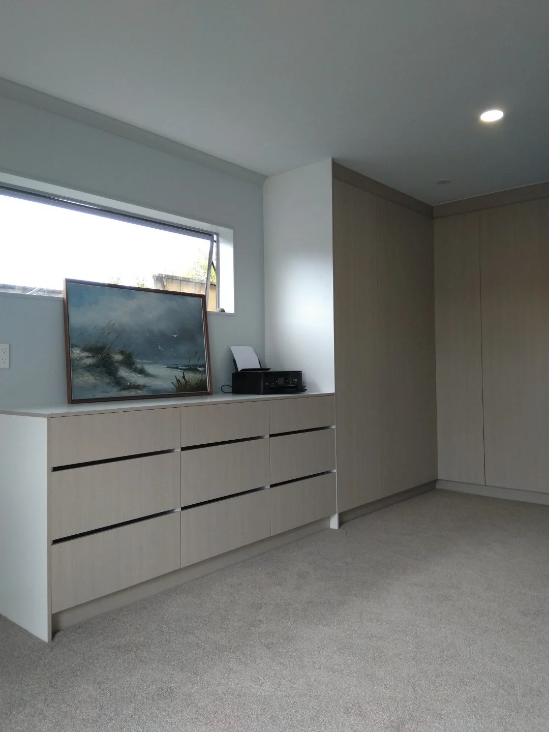 Empty modern room with light-colored wood storage units, a small window, a framed painting leaning on the wall, and a printer.