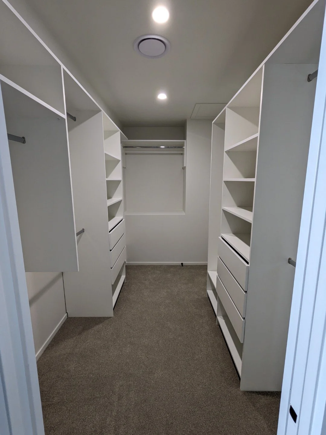 Empty walk-in closet with white built-in shelves and drawers, gray carpet, and ceiling lights.