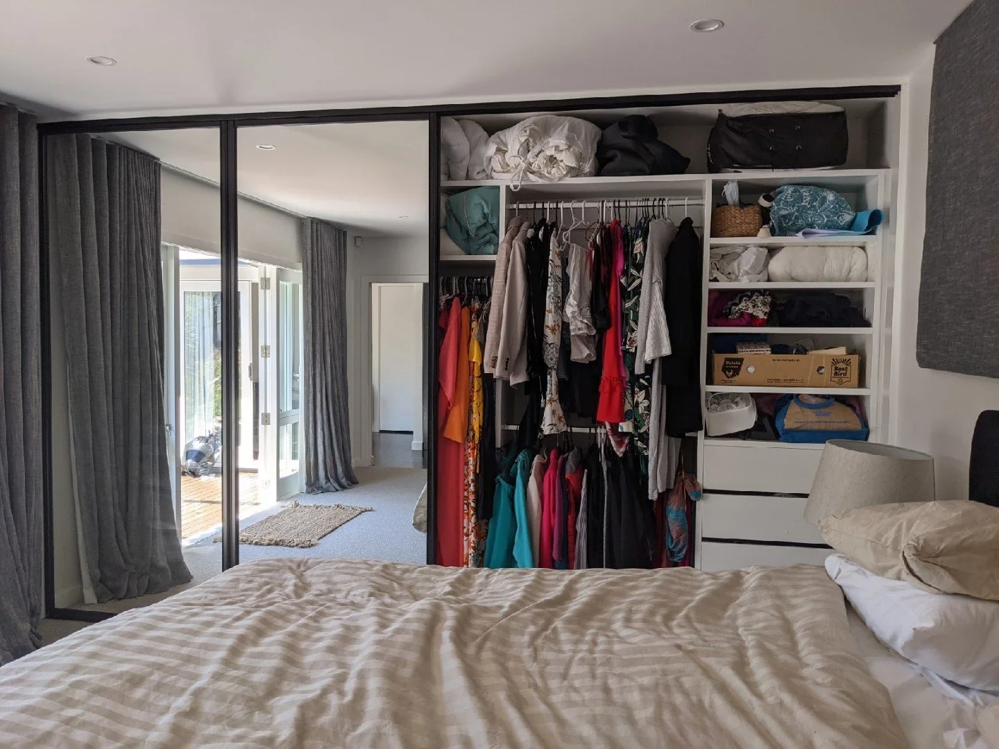 A bedroom with a bed, bedside lamp, and an open closet with hanging and folded clothes, bags, and storage boxes, adjacent to a mirrored sliding door that opens to a patio area.
