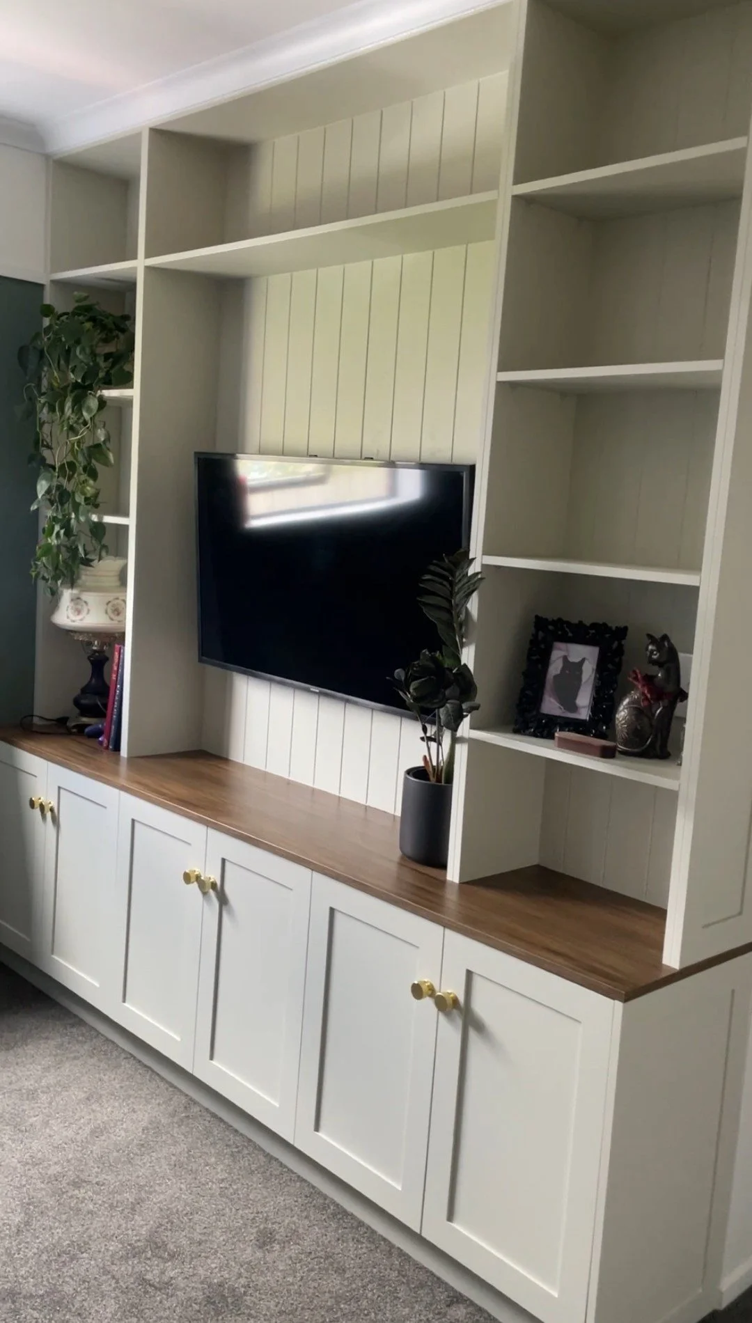 White entertainment unit with shelves and cabinet doors, a mounted TV, and decorative items including plants and photo frames.