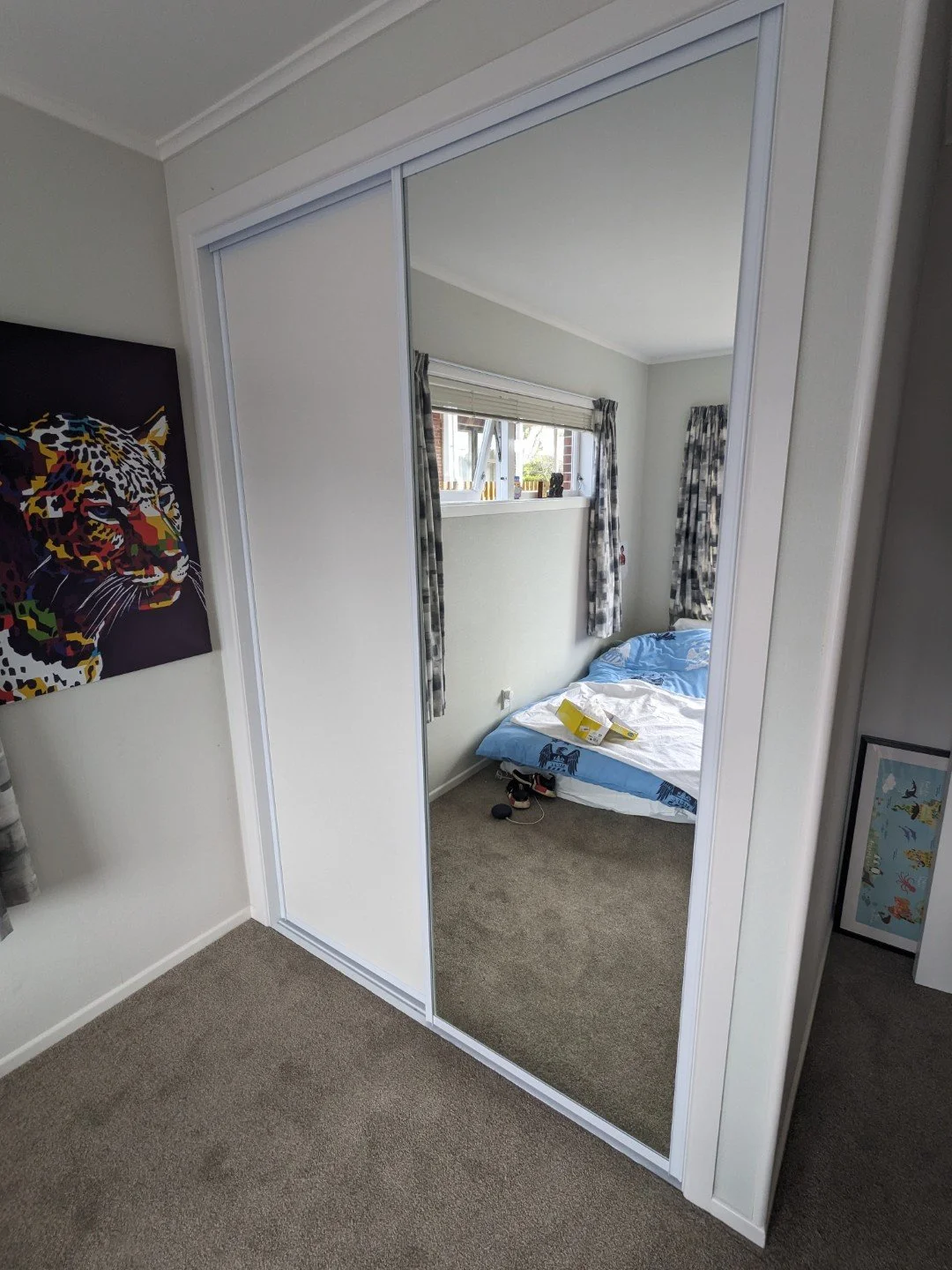 A bedroom with a mirrored sliding door wardrobe, a window with curtains, and a bed with blue and white bedding. Clothes and slippers are on the floor next to the bed.