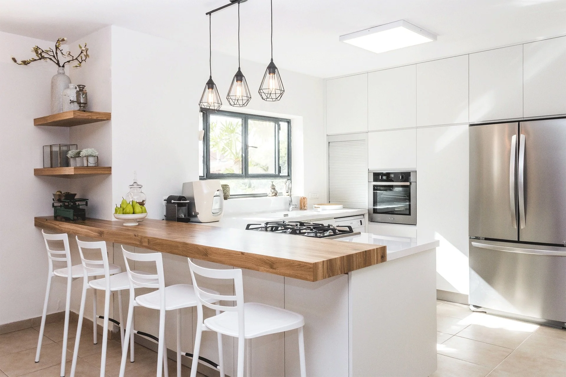 Modern kitchen with white cabinets, stainless steel refrigerator, oven, and wooden countertop with bar stools. Decorative shelves with vases and plants, and a window letting in natural light.