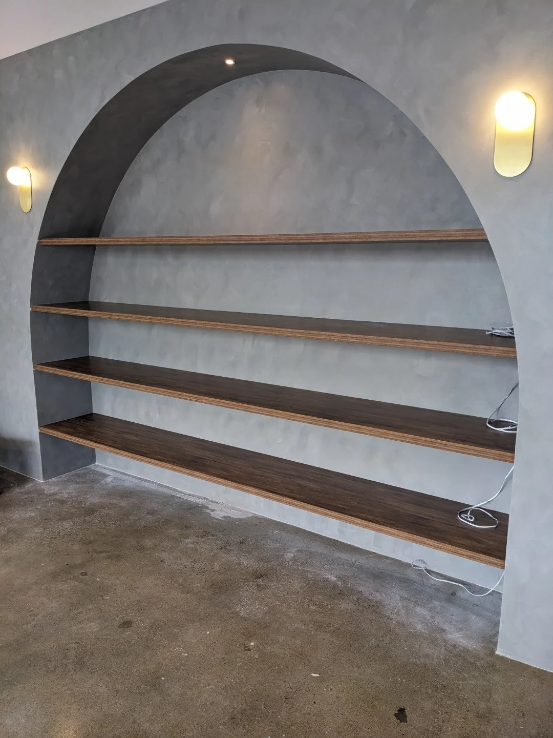 Built-in floating wooden shelves with an arched cutout design on a concrete floor, with wall-mounted lights on either side, and electrical cords on the shelves for LED lighting.