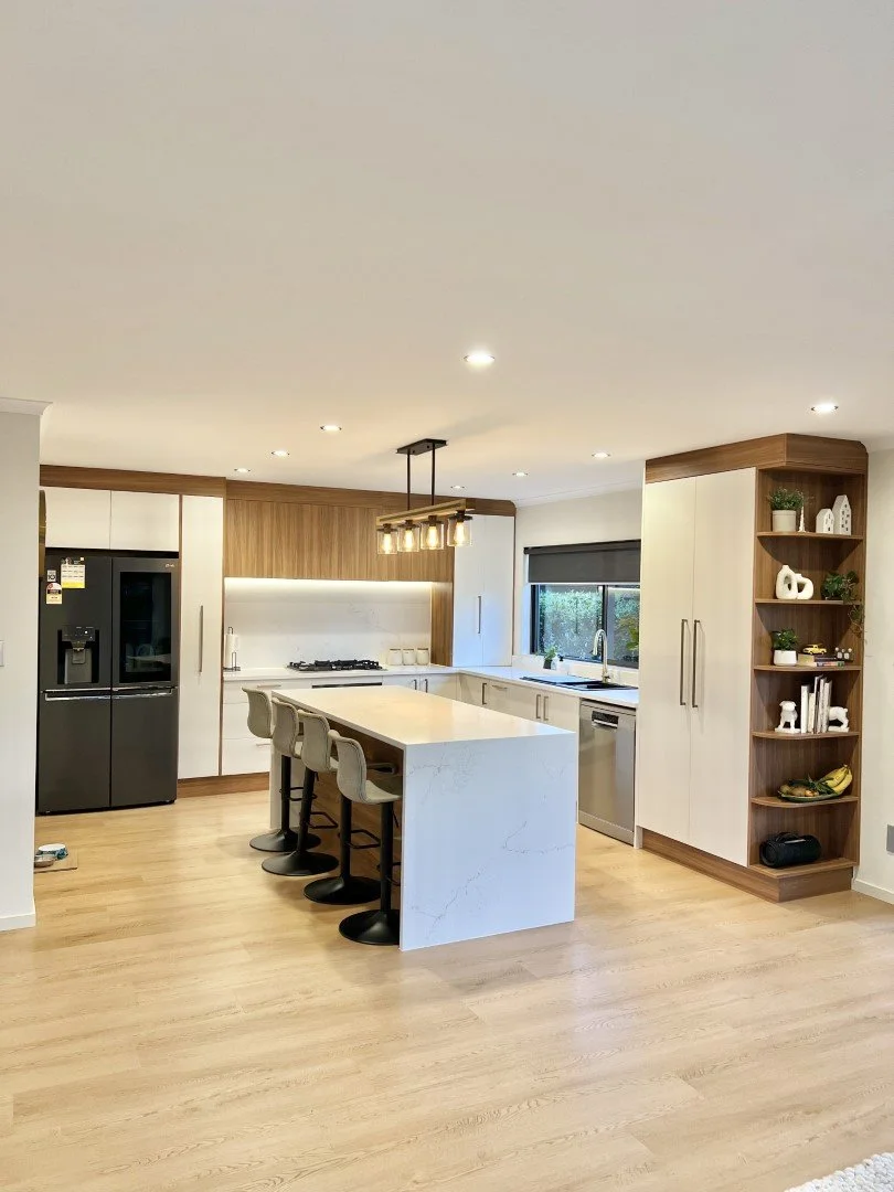 Modern kitchen with white and wood cabinetry, marble island with four bar stools, black refrigerator, and stainless steel appliances, illuminated by recessed ceiling lights and a chandelier, with a window over the sink and open shelving with decorati