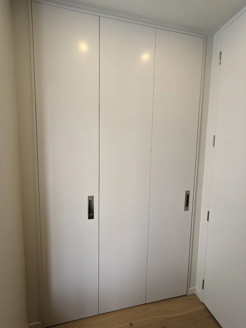 White triple bi-fold doors, fixed door on the right side, situated in a laundry room with light-colored walls and wooden flooring.